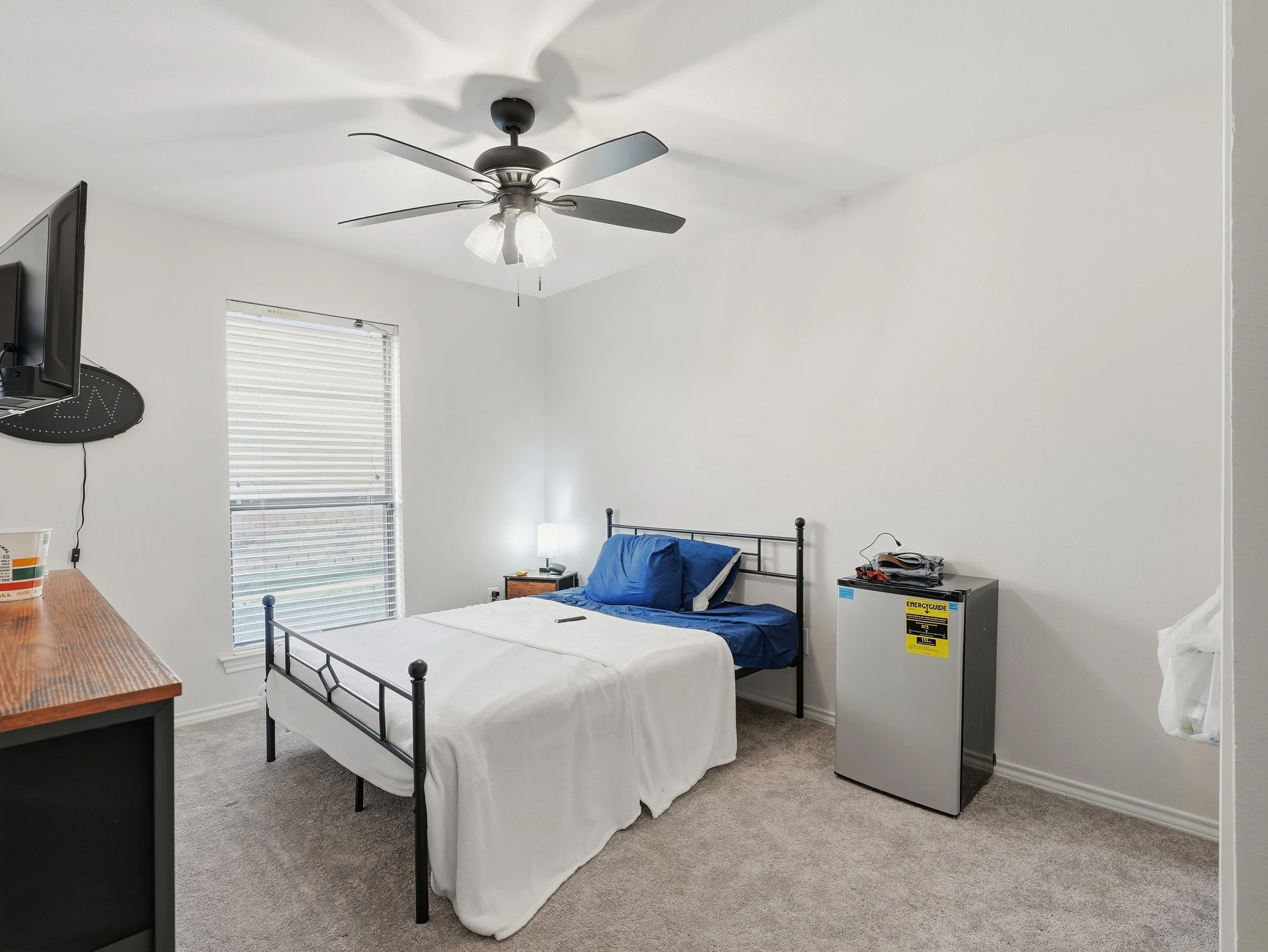 A simple bedroom with a bed, window, small nightstand with lamp, mini refrigerator, and a TV mounted on the wall.