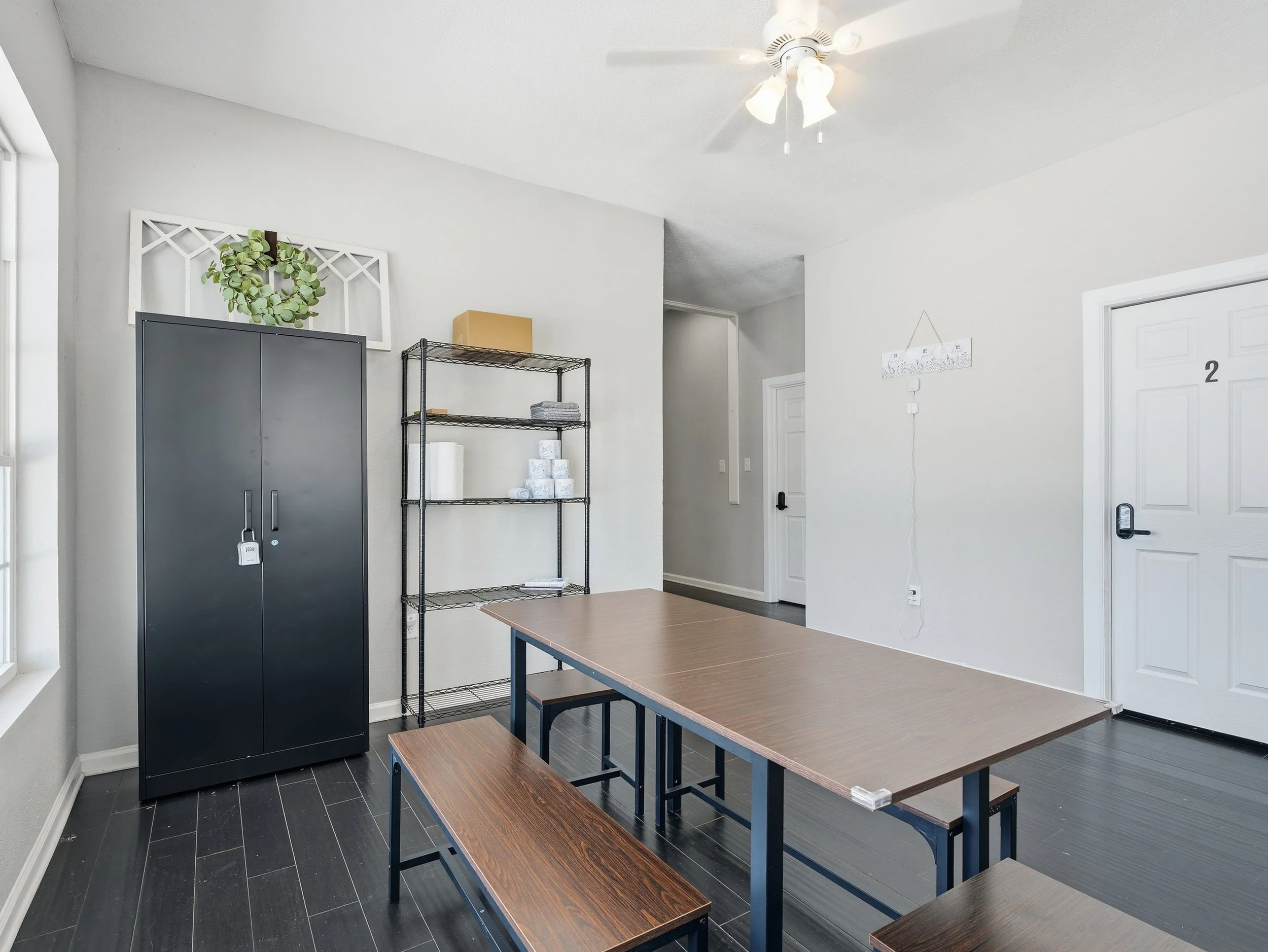 Room with white walls, a dark gray cabinet with a lock, a black metal shelving unit with tissues and a box, a wooden table with matching benches, a ceiling fan, and a white door with the number 2.