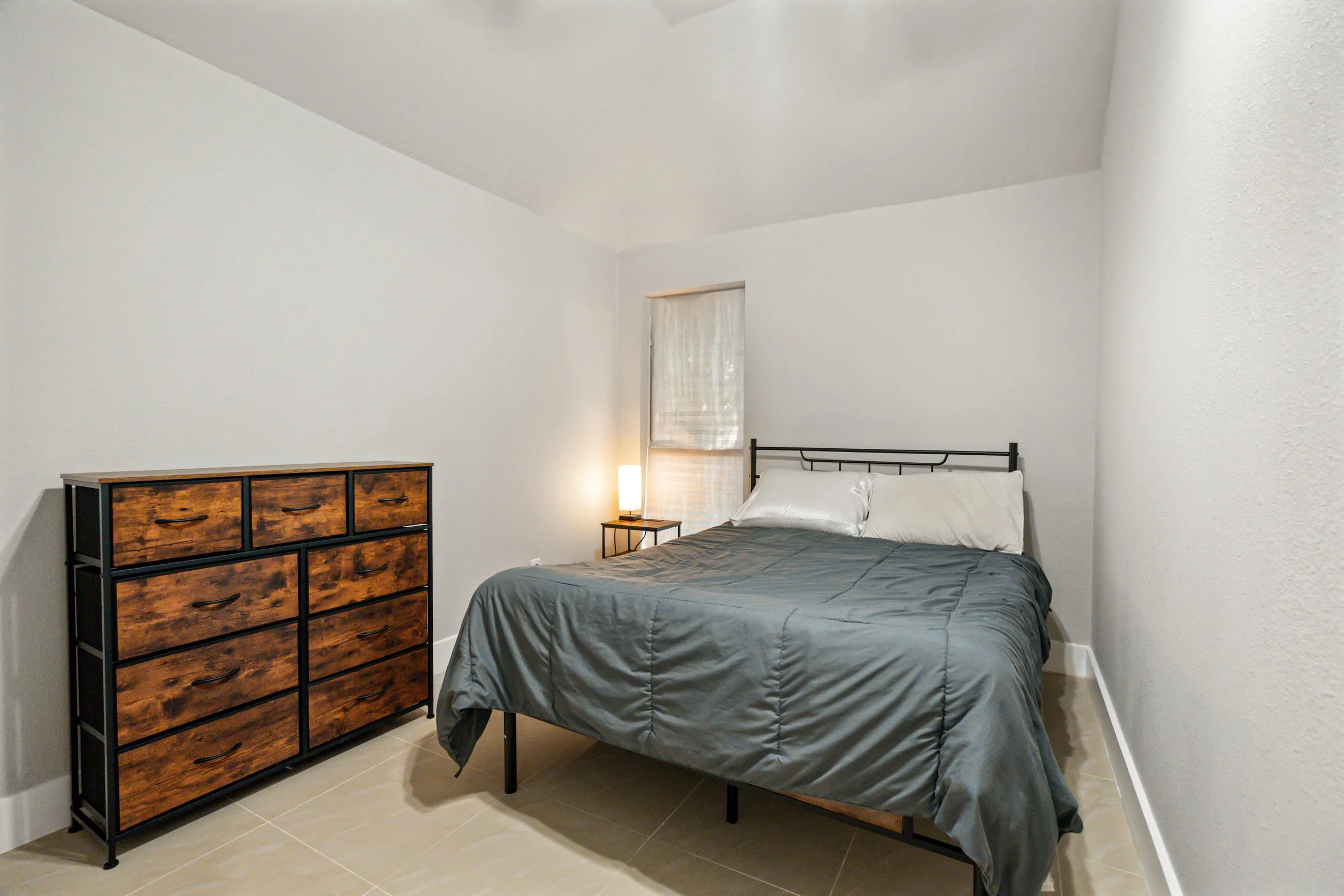 Minimalist bedroom with a bed, gray bedding, white pillows, a nightstand with a lamp, a wooden dresser, and a window with white blinds.