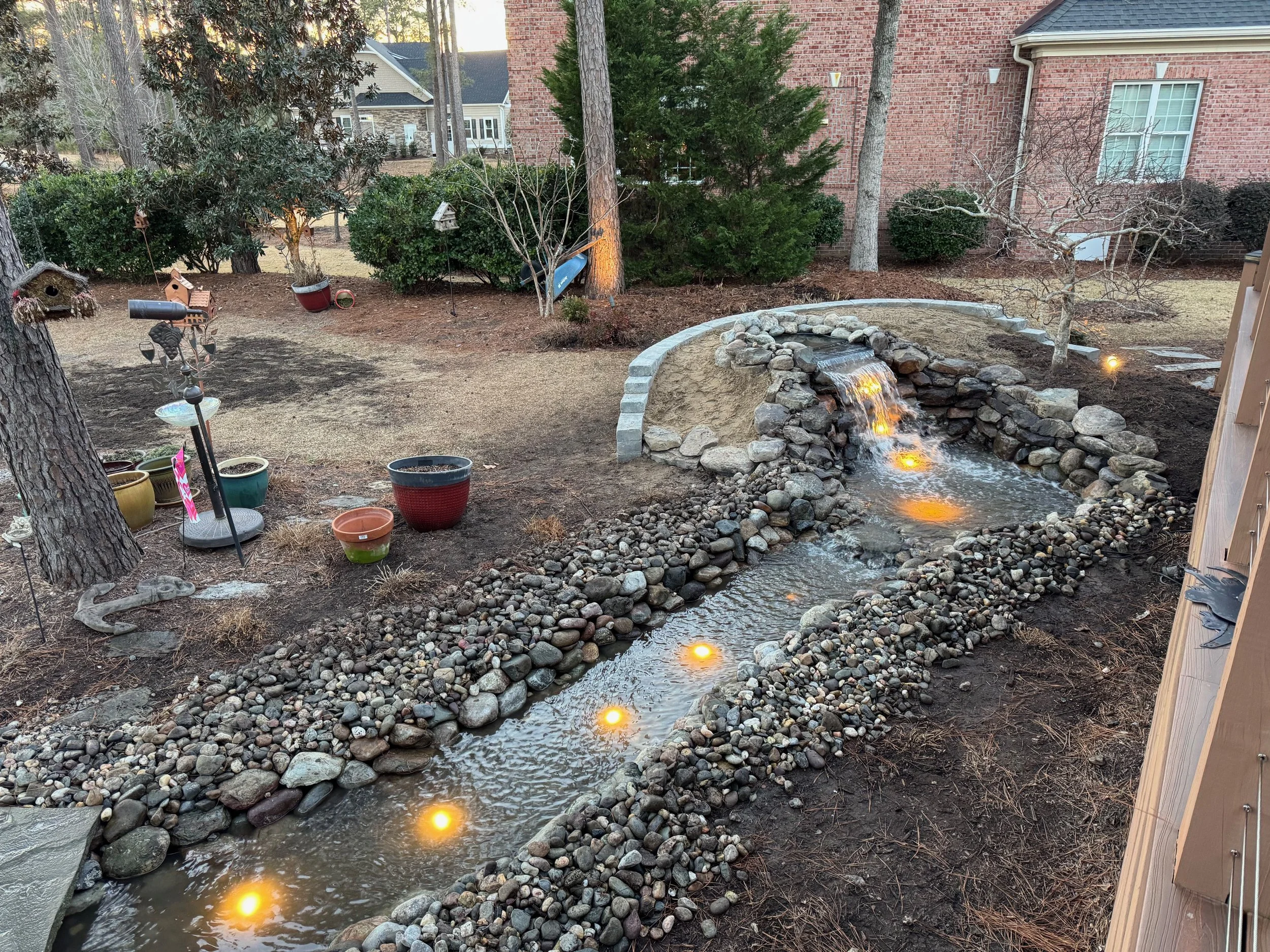 Backyard landscape with a small waterfall and stream illuminated by embedded lights, surrounded by rocks and mulch. Decorative items, pots, and a birdhouse are visible along with trees and neighboring houses.
