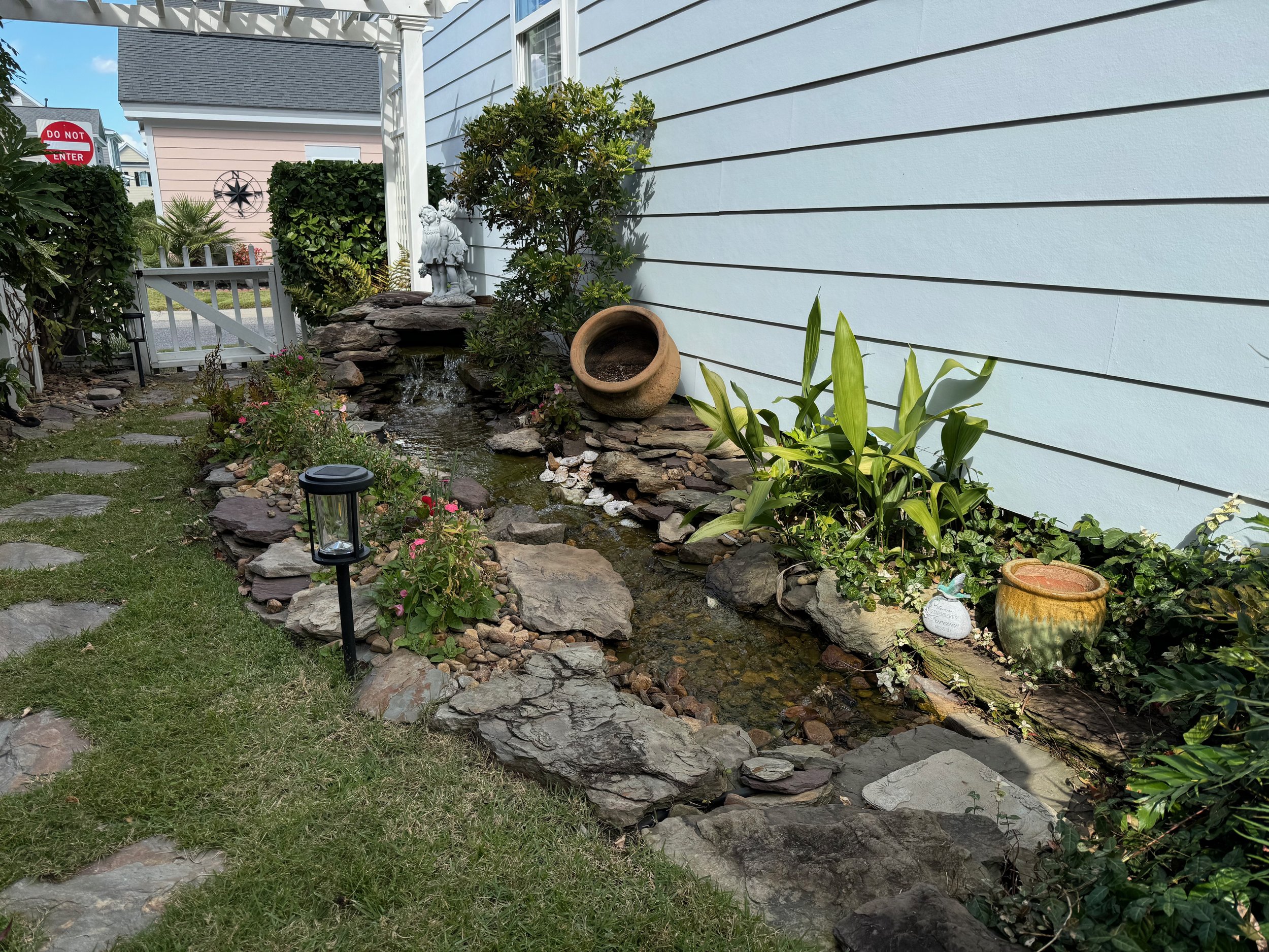 A small backyard garden with a stone-lined pond, decorative pots, lush greenery, blooming flowers, and a white house siding wall.