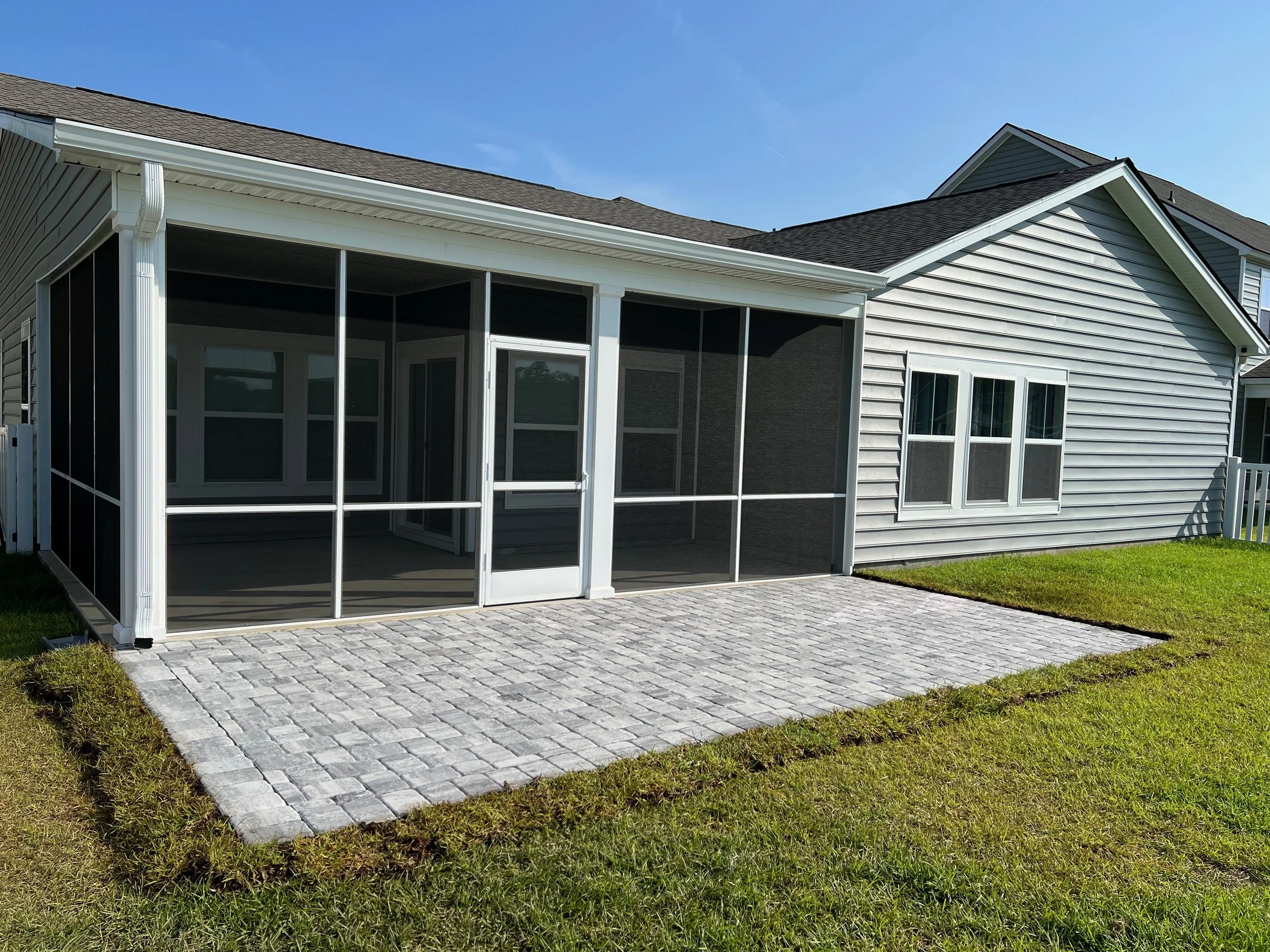 Backyard patio with gray brick pavers and a screened-in porch attached to a house with light gray siding, white trim, and multiple windows. The yard has green grass, and the sky is clear and blue.