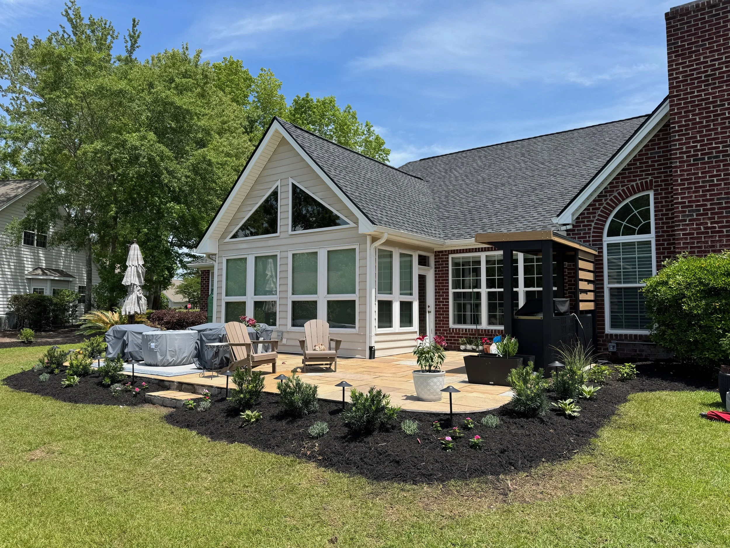 Backyard patio with Adirondack chairs, potted plants, grill, and a sitting area, surrounded by flower beds and a well-maintained lawn beside a house with brick and siding, under a blue sky with some clouds.