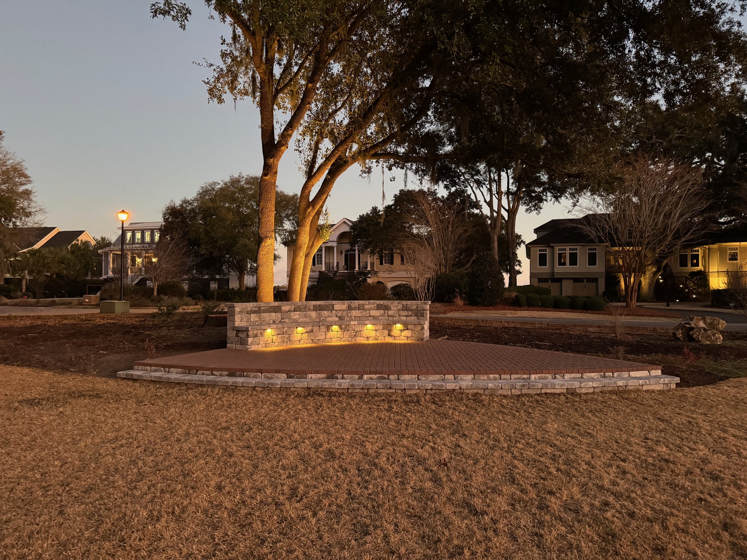 A park at dusk with a small brick stage with three square yellow lights on a low brick wall, trees, houses in the background, and a lamppost on the left side.