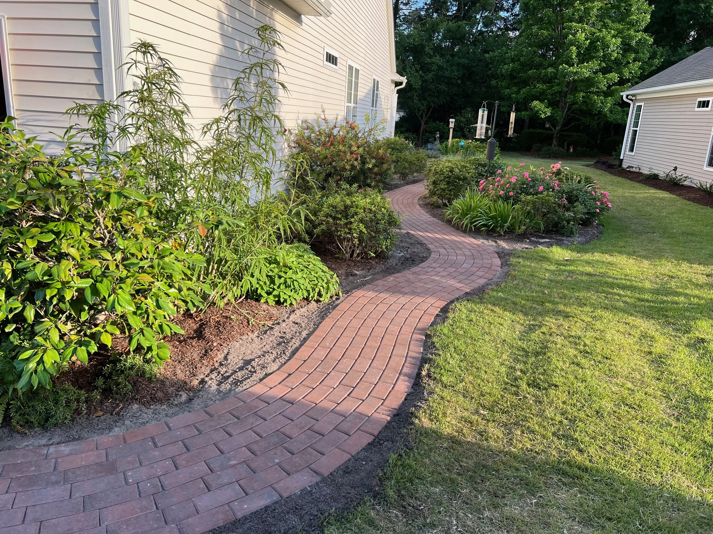 Curved brick pathway through a well-kept garden with green grass, bushes, pink flowers, and garden lights in a residential backyard.