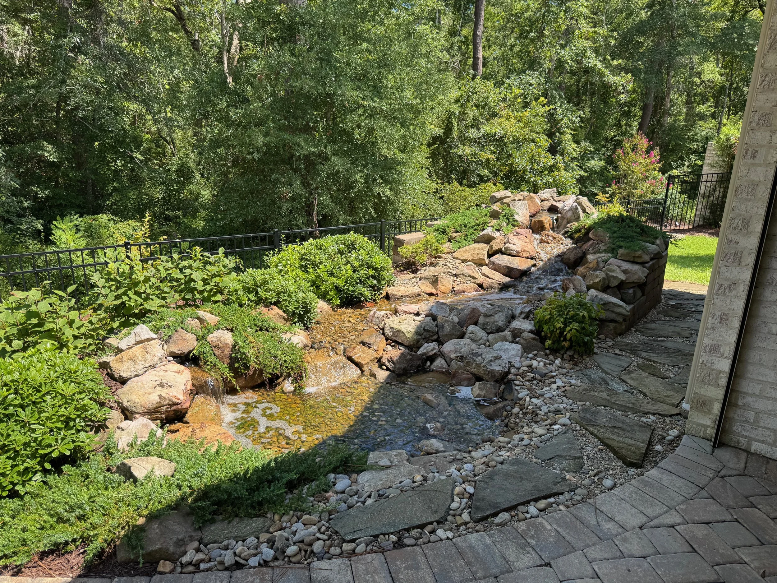 Backyard garden with a small waterfall, rocks, and lush greenery surrounded by a black metal fence and a brick house wall.