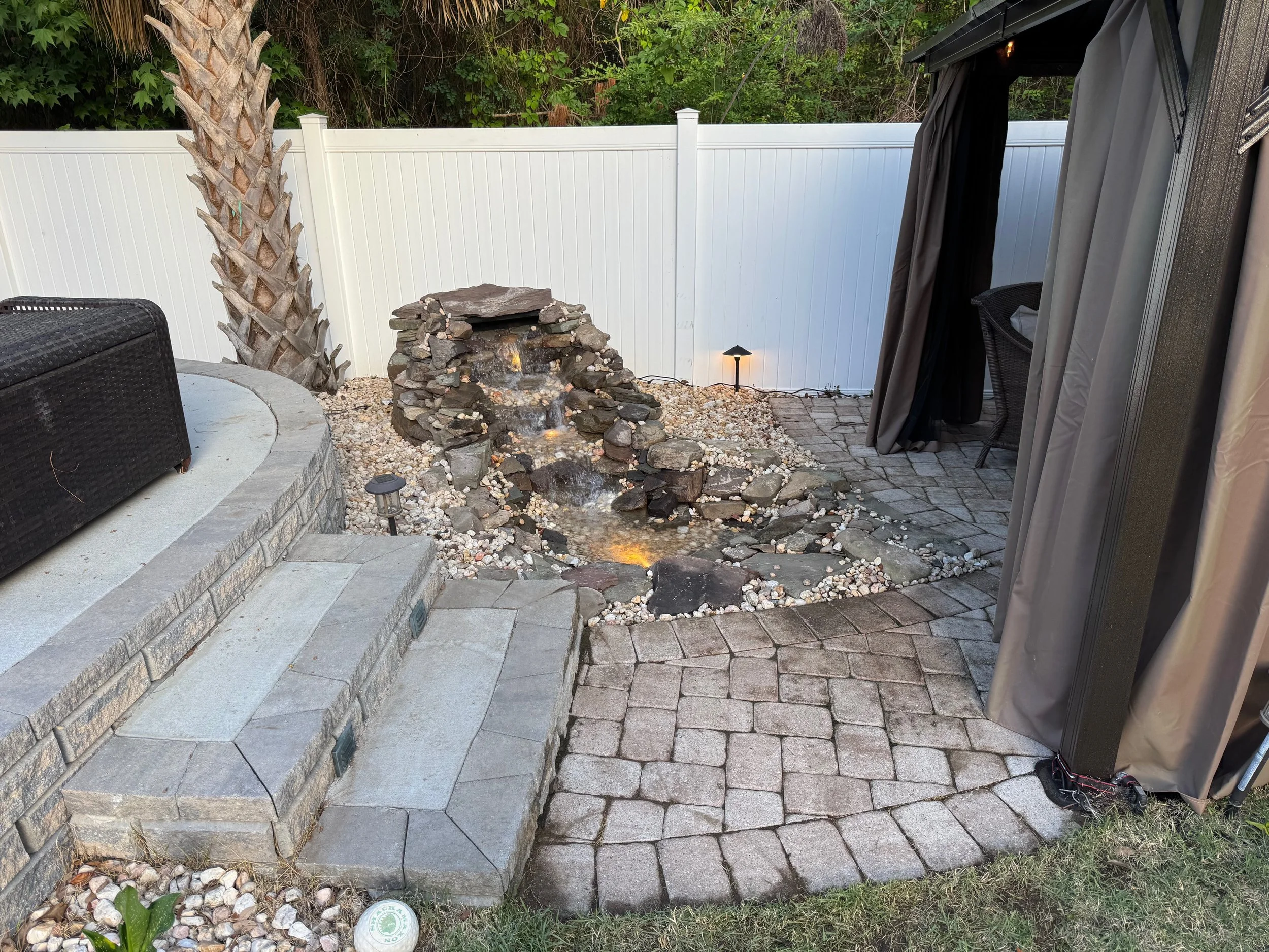 Backyard patio with a small waterfall feature, paver stones, a white privacy fence, patio furniture, and outdoor lighting.