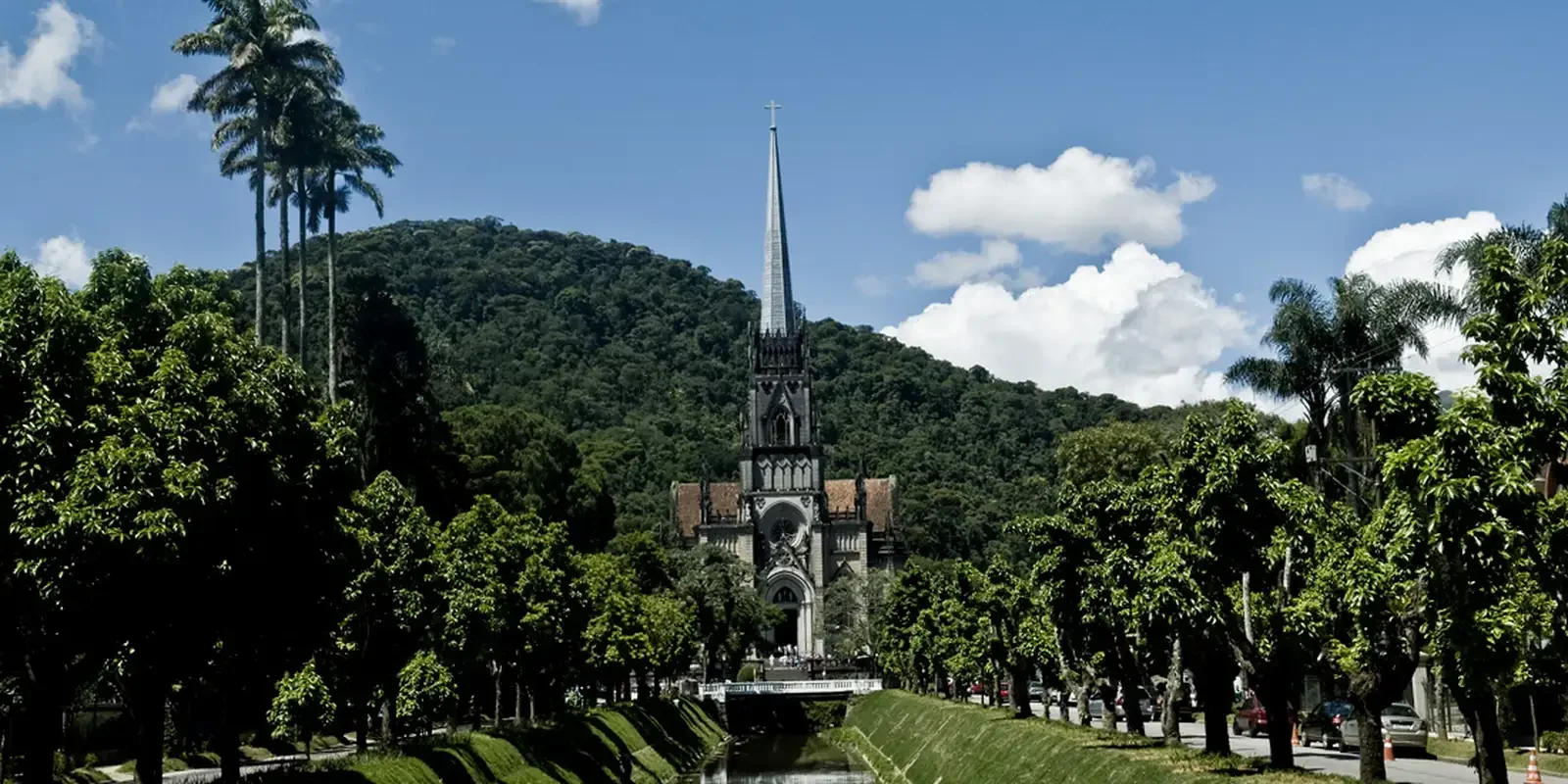 Igreja em estilo gótico com torre alta e detalhes ornamentais, cercada por árvores verdes e um rio na frente, com montanha ao fundo e céu azul com algumas nuvens.