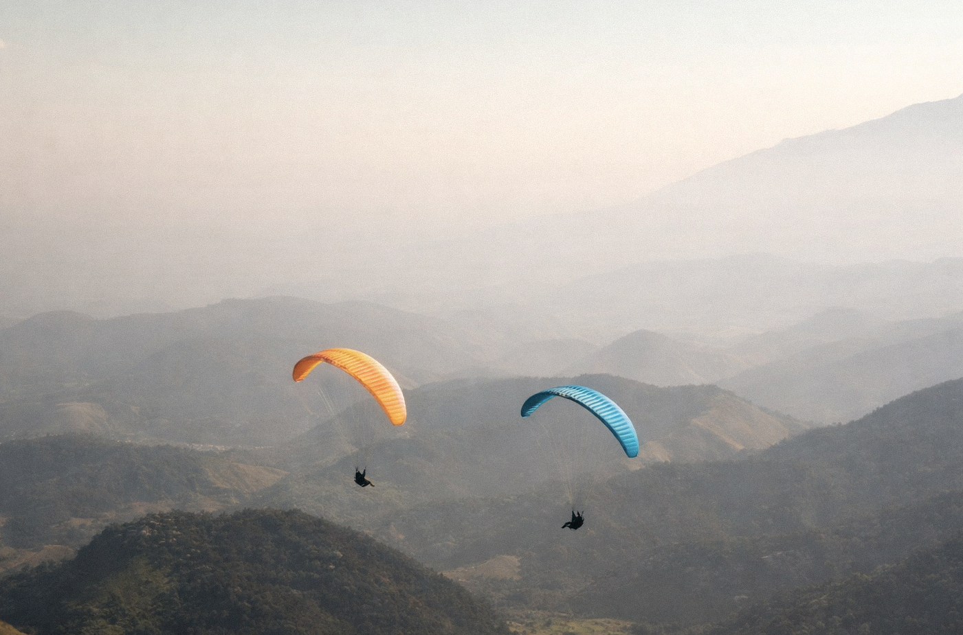 Duas pessoas praticando parapente sobre paisagem montanhosa ao pôr do sol, com céu claro e nublado ao fundo.