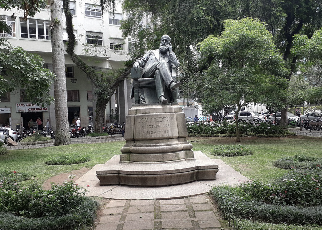 Estátua de Dom Pedro II sentado em um banco no parque, rodeado de árvores e plantas, com edifícios e carros ao fundo.