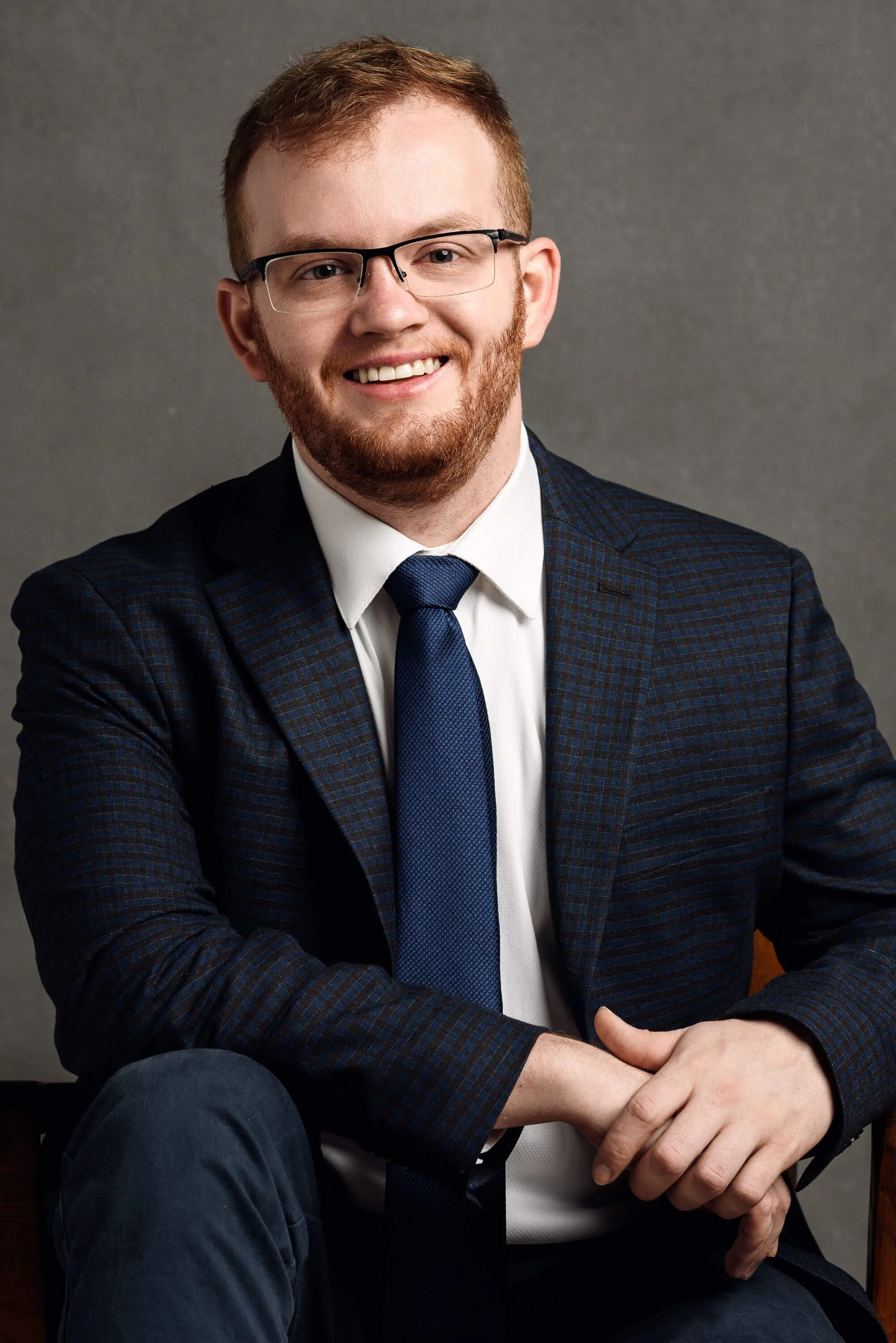 A young man with red hair, glasses, and a beard, wearing a navy plaid blazer, white shirt, and navy tie, sitting against a gray background and smiling.
