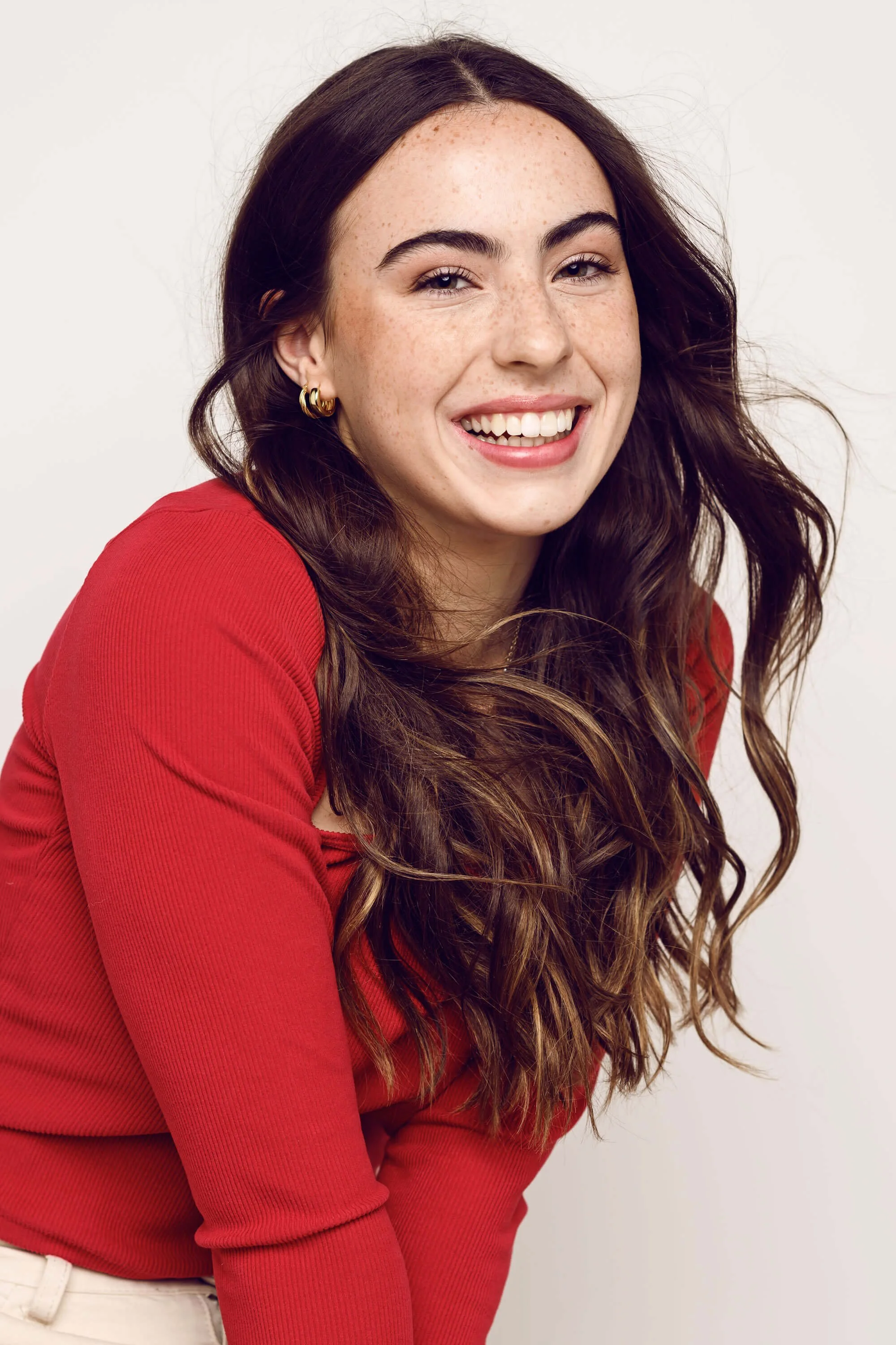 A young woman with long wavy brown hair, freckles, and gold hoop earrings, smiling and wearing a red long-sleeve top against a light background.