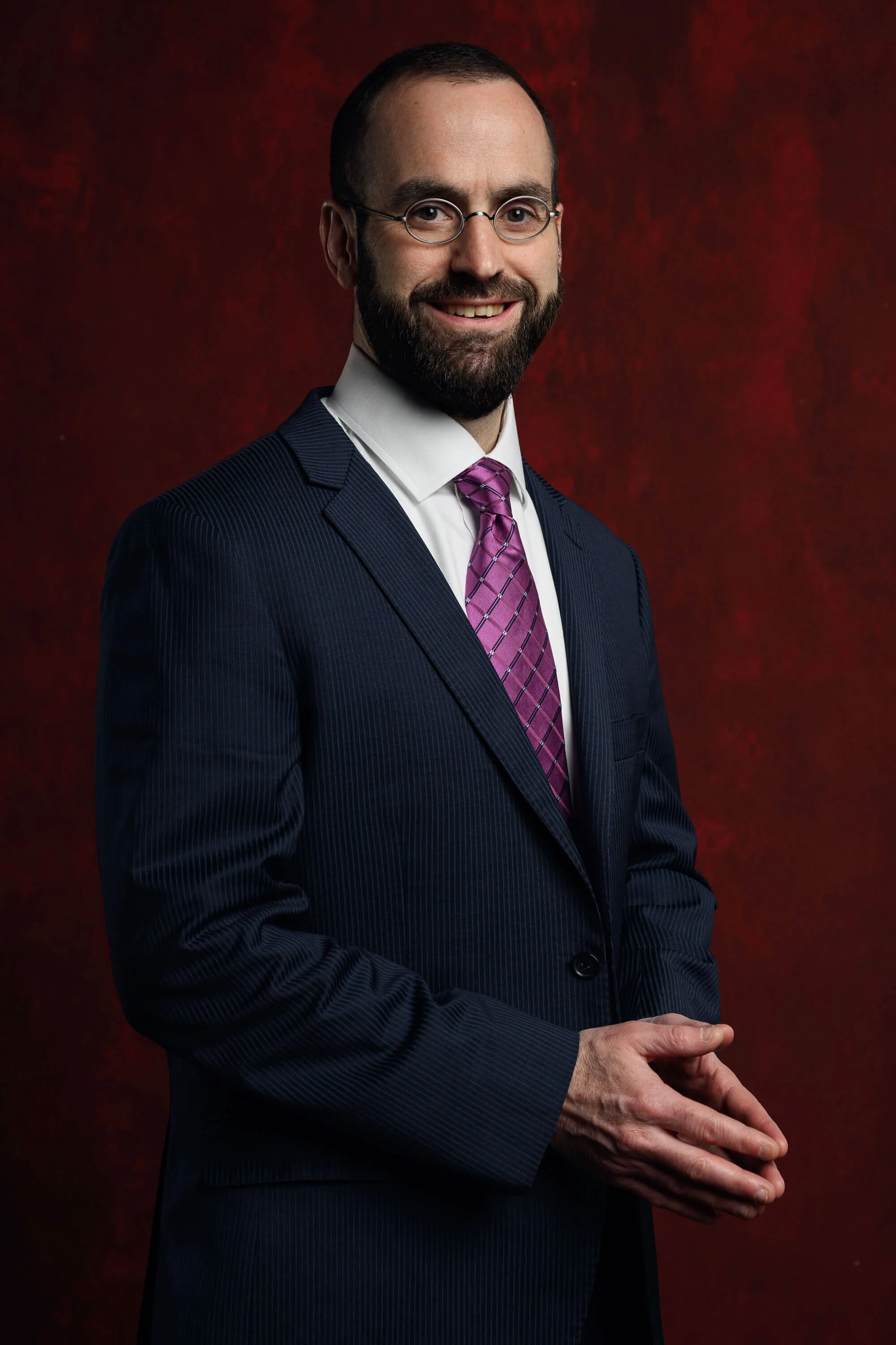 A man with glasses and a beard in a navy pinstripe suit, white shirt, and pink tie against a dark red background.