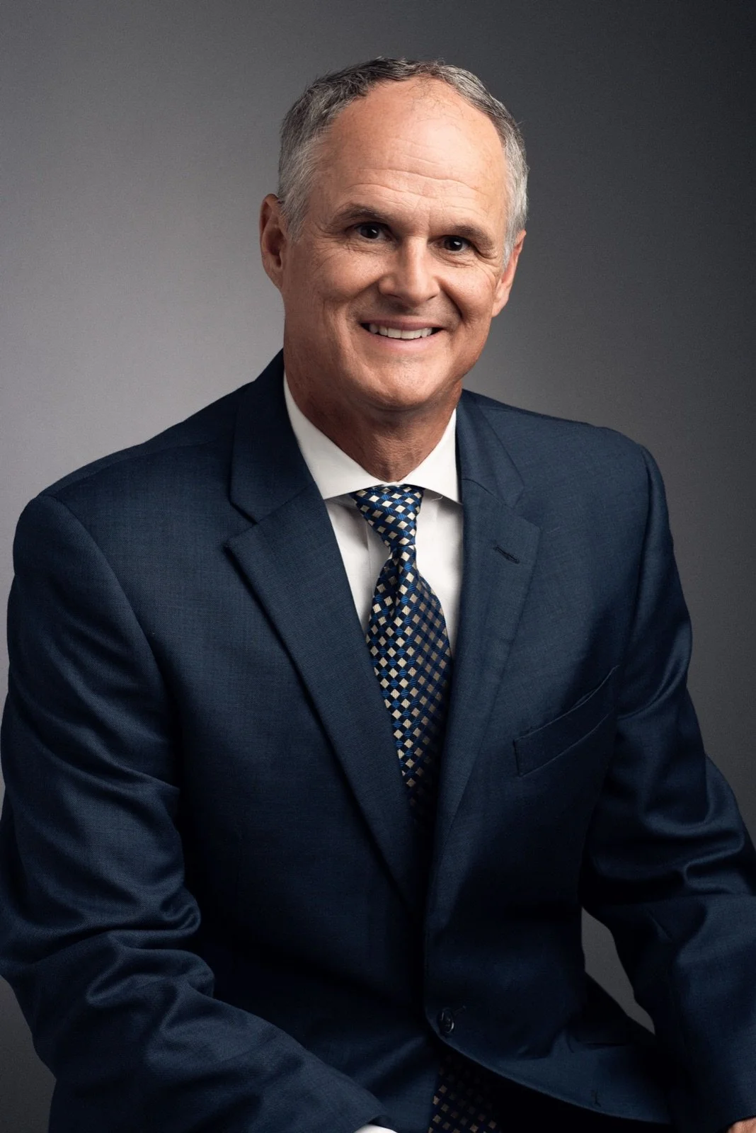 A middle-aged man with short gray hair, wearing a dark blue suit, white shirt, and multicolored checkered tie, smiling at the camera against a neutral gray background.