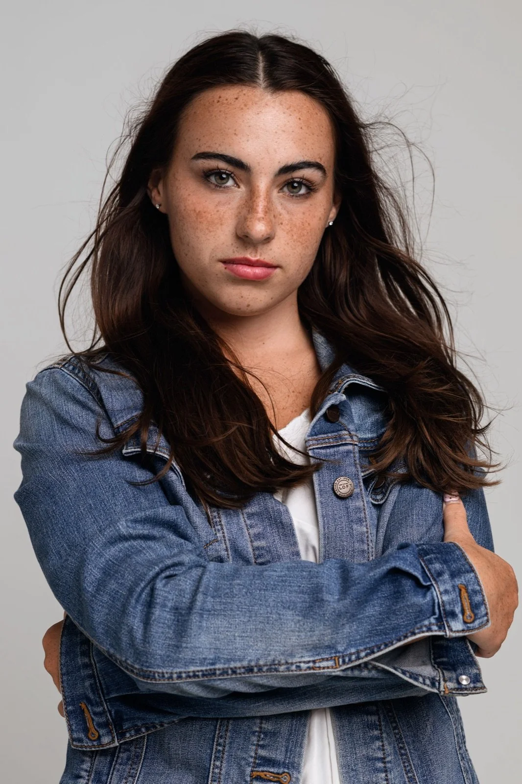 A young woman with long brown hair, freckled face, wearing a denim jacket, crossing her arms and looking directly at the camera.