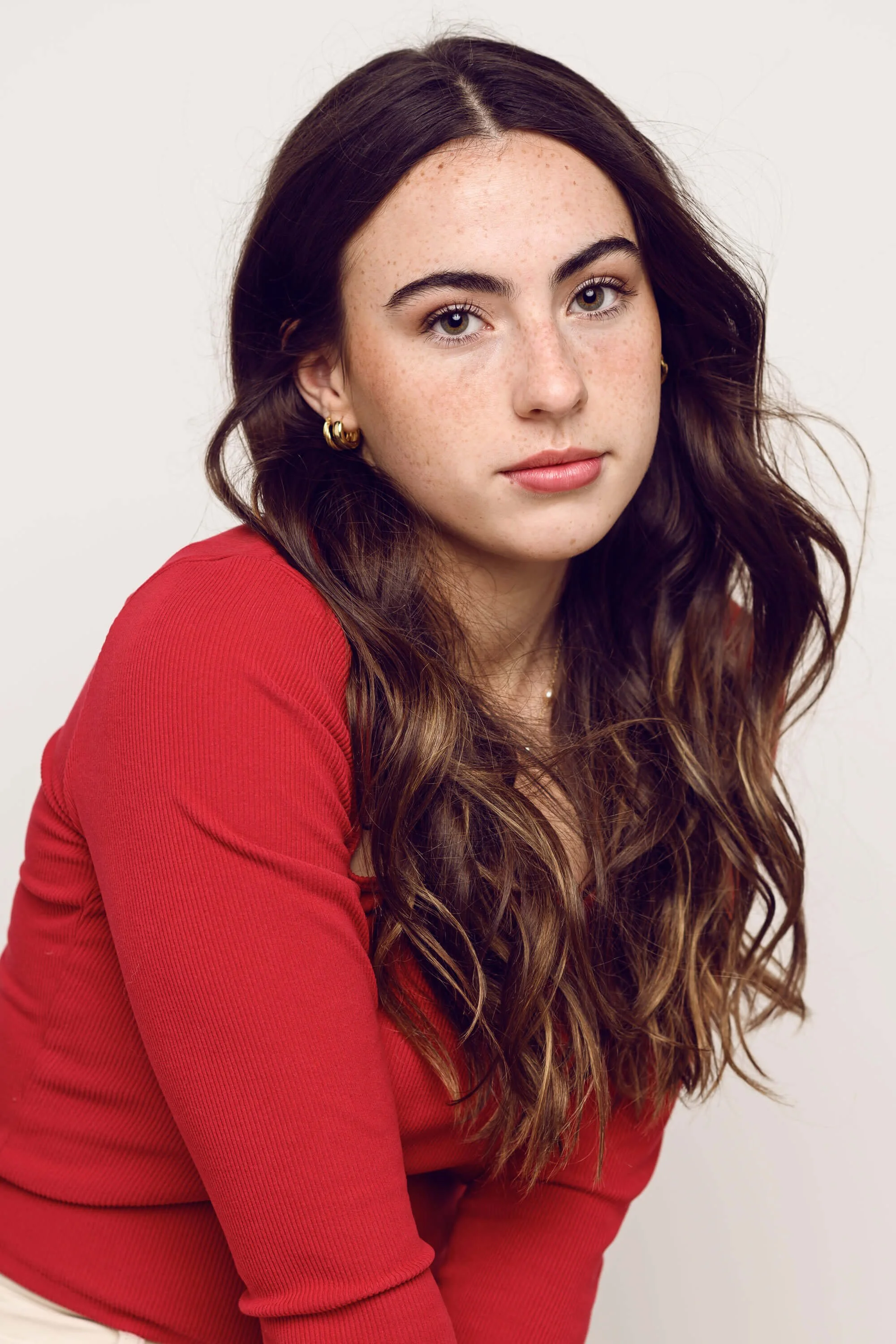 A young woman with long wavy brown hair and light freckles, wearing hoop earrings and a red long-sleeve top, seated against a plain white background.