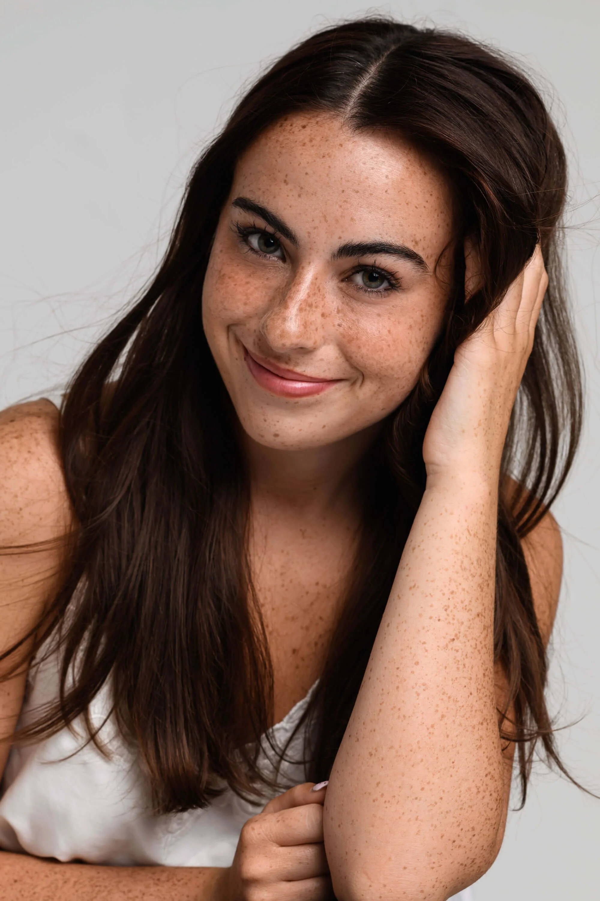Close-up portrait of a young woman with brown hair and freckles, smiling and resting her head on her hand against a plain background.
