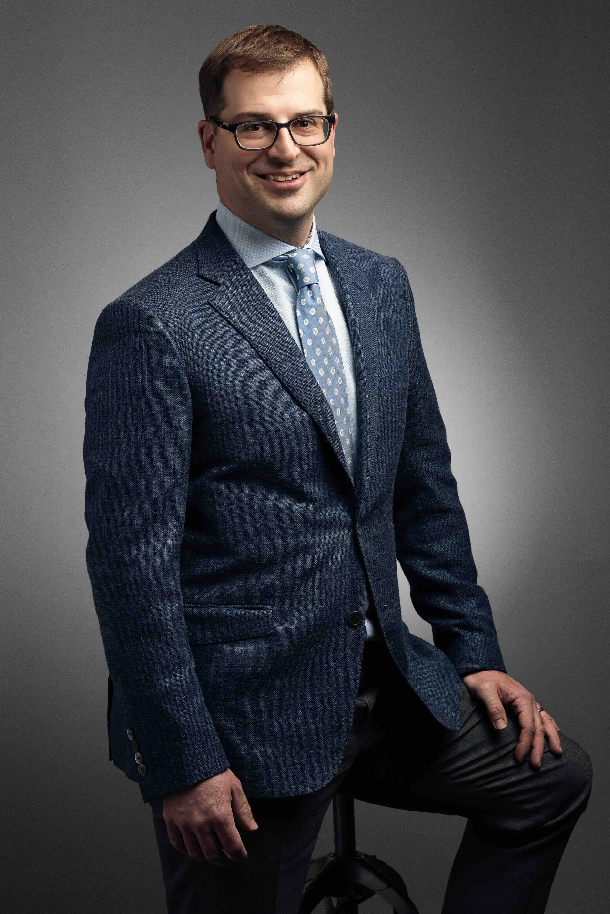 A man in a dark blue suit with a light blue shirt and matching polka dot tie, wearing glasses, sitting on a stool against a plain gray background.