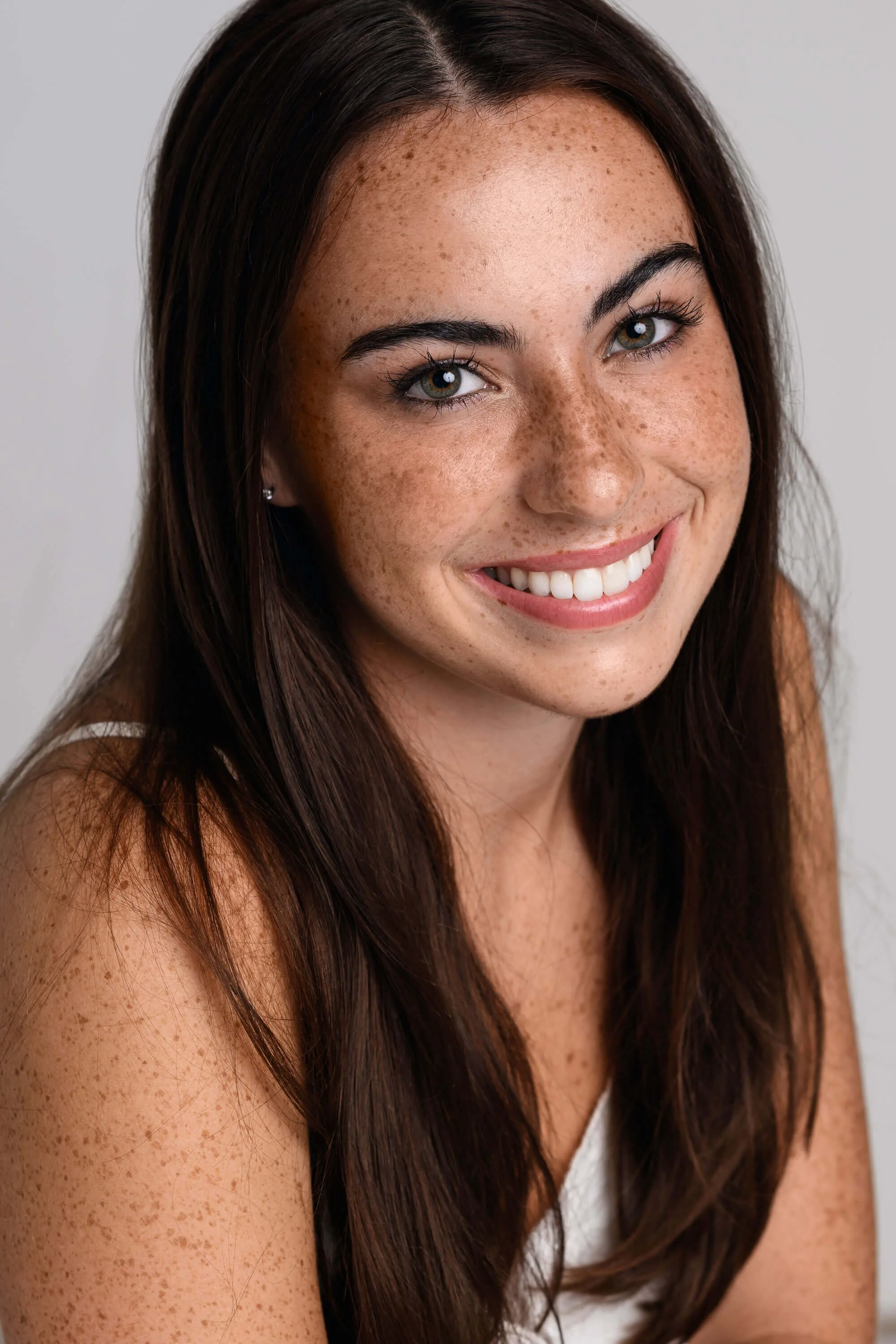 Close-up portrait of a smiling woman with long brown hair and freckles, wearing a white top.