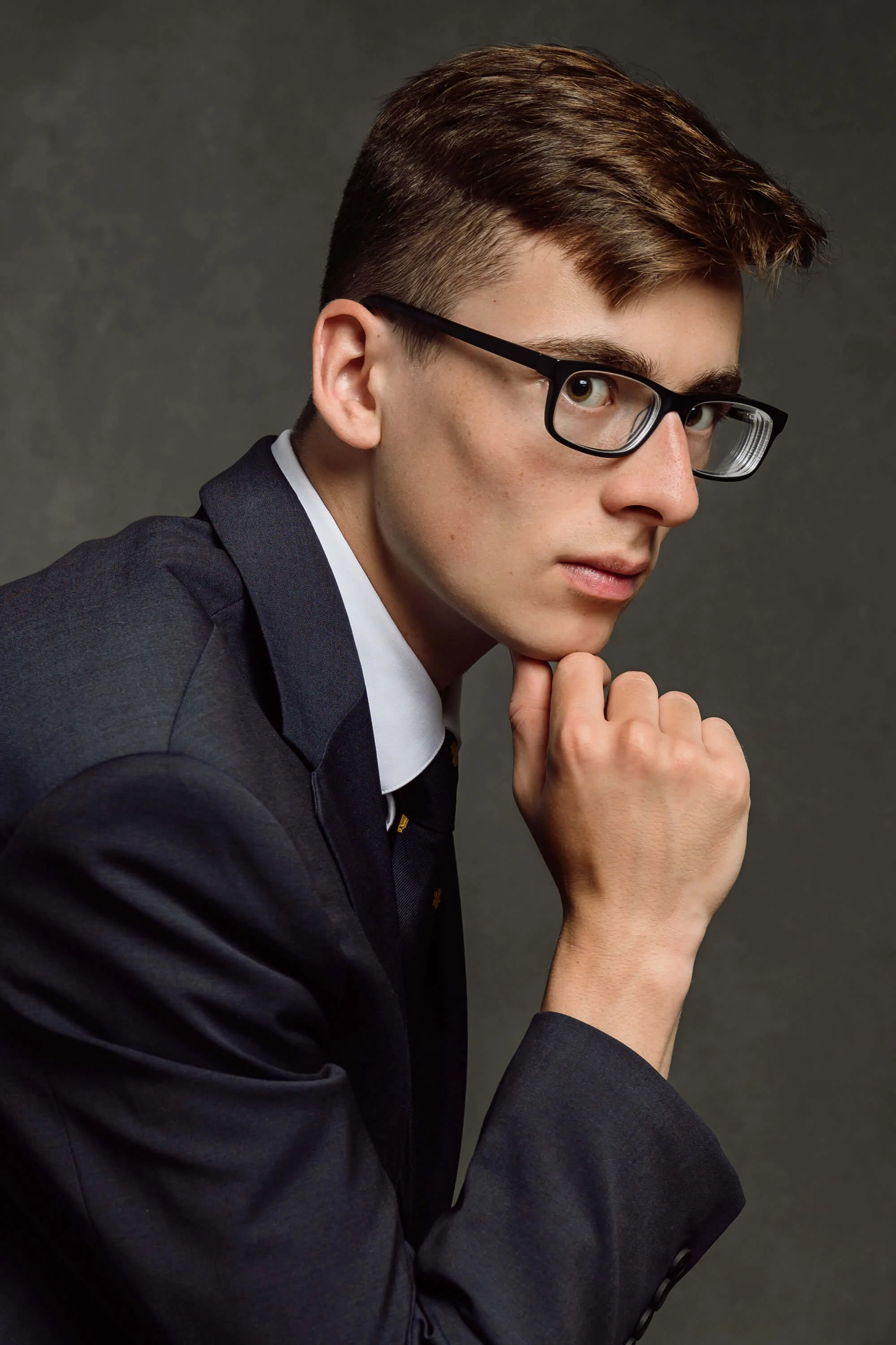 A young man with glasses and short brown hair, wearing a dark suit and tie, looking thoughtfully at the camera with his hand resting on his chin.