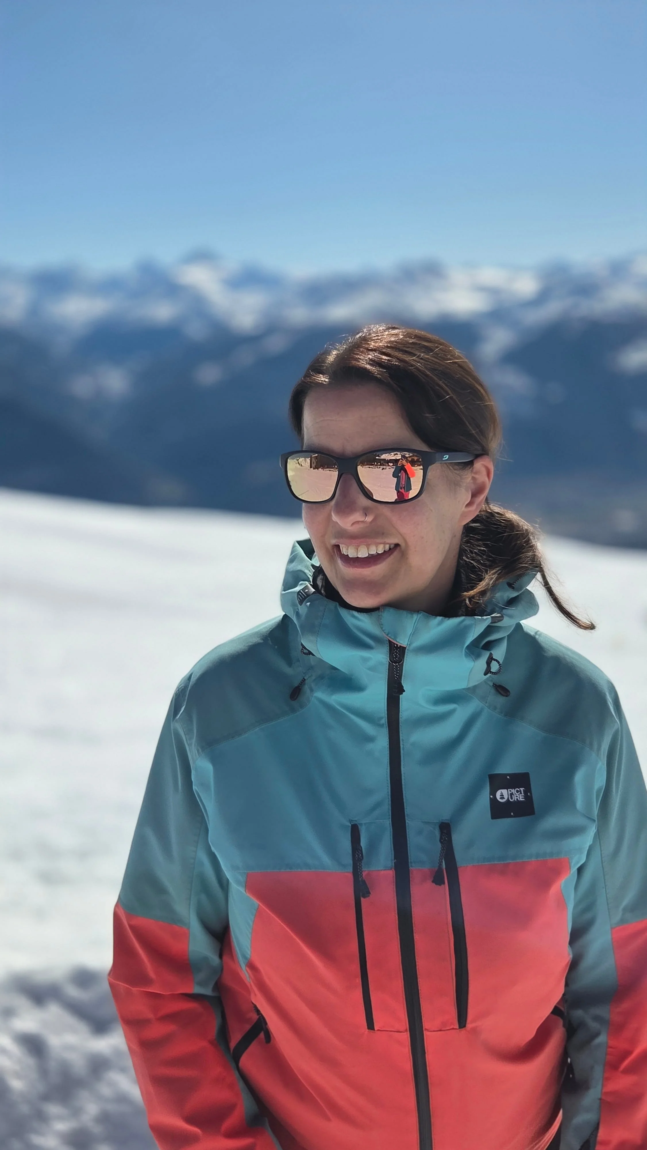 Eine lachende Frau in einer bunten Skijacke und Sonnenbrille in verschneiter Berglandschaft bei Sonnenschein.