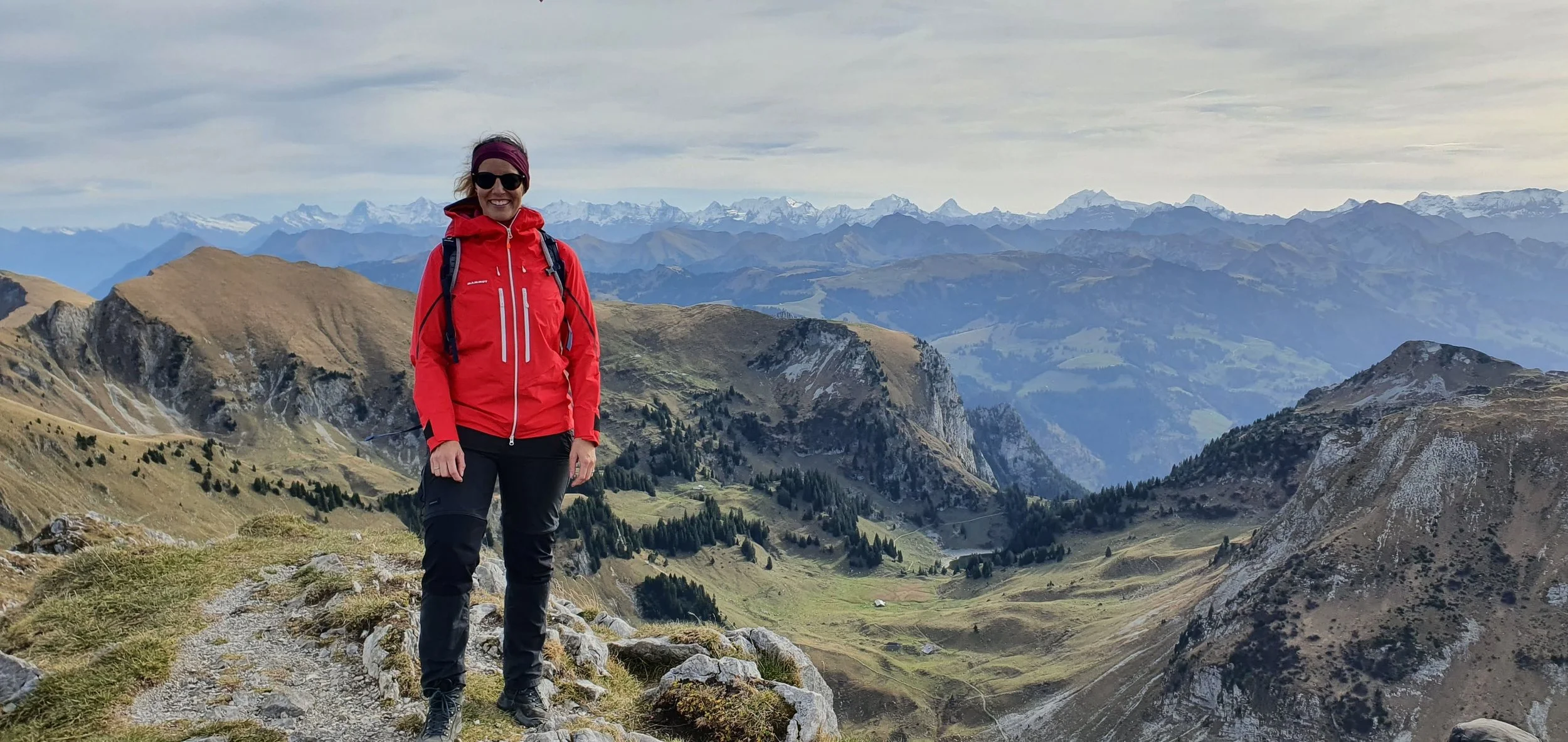 Frau in roter Jacke und schwarzen Hosen beim Wandern in den Bergen mit Blick auf eine bergige Landschaft und schneebedeckte Gipfel im Hintergrund.
