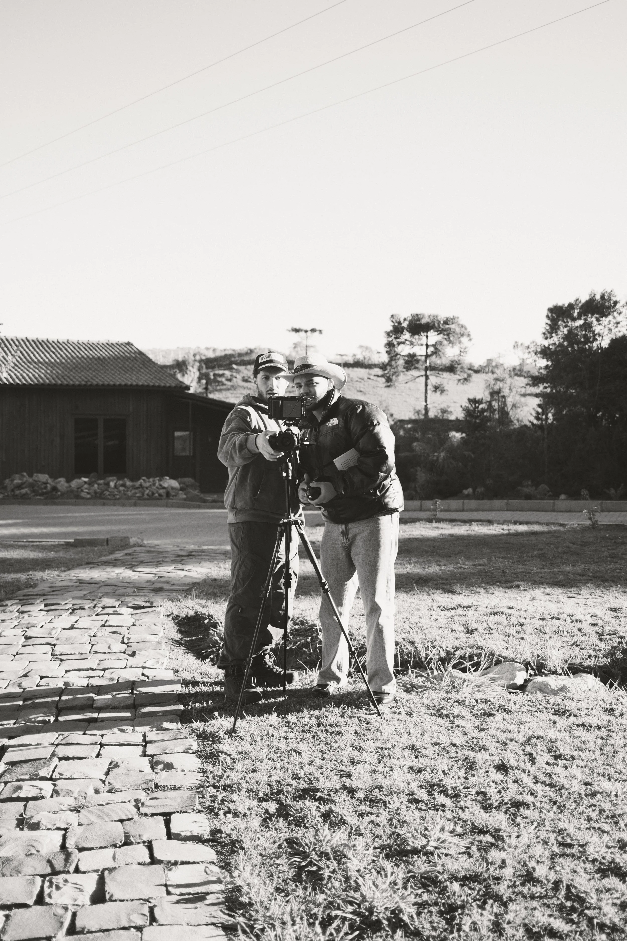 Dois homens ajustando uma câmera em um tripé ao ar livre, em um ambiente rural, com uma casa de madeira ao fundo, árvores e um céu claro.
