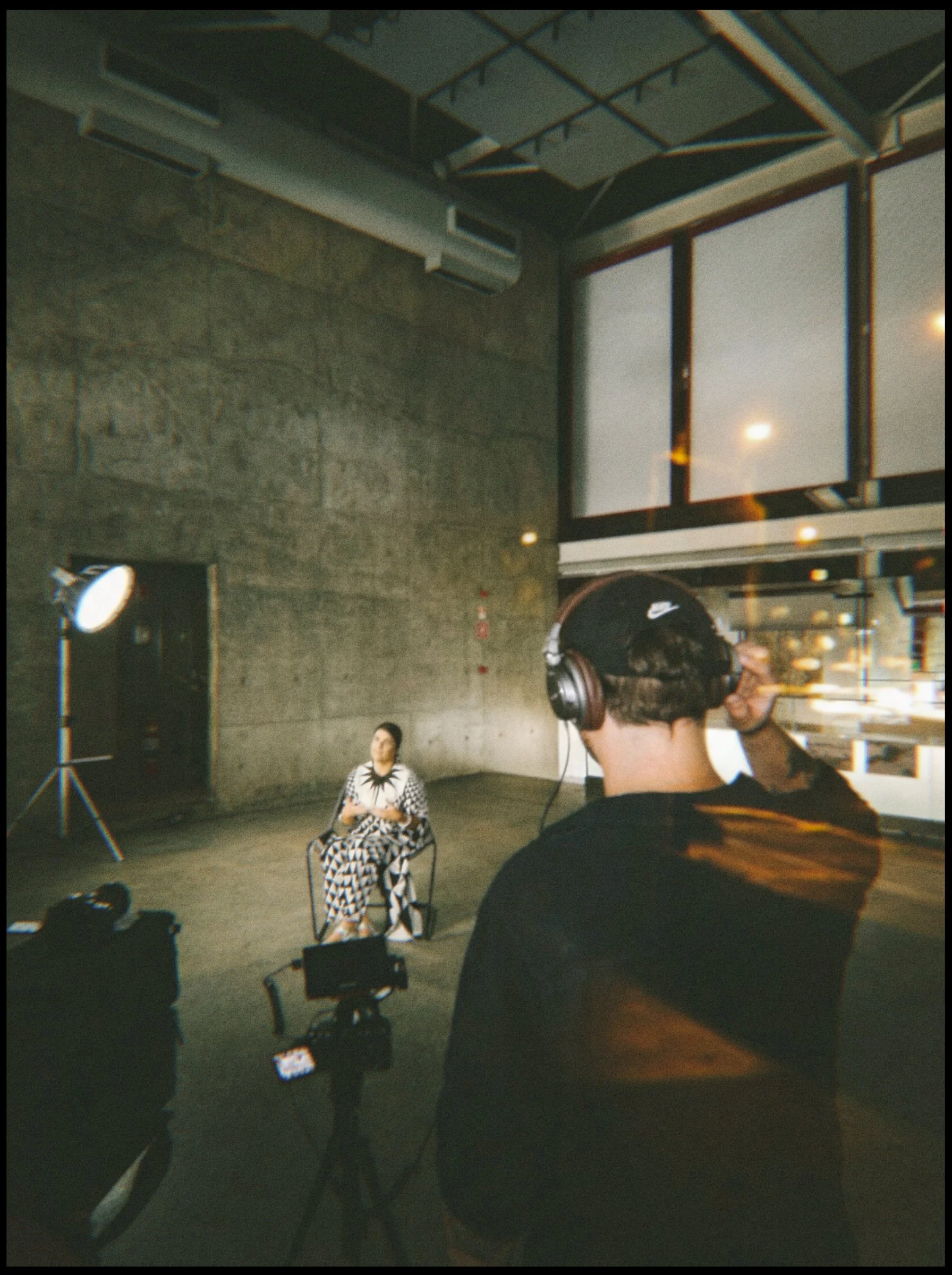 Pessoa com fones de ouvido grava vídeo de uma mulher sentada em uma cadeira em um estúdio com iluminação profissional, paredes de cimento e janelas grandes.