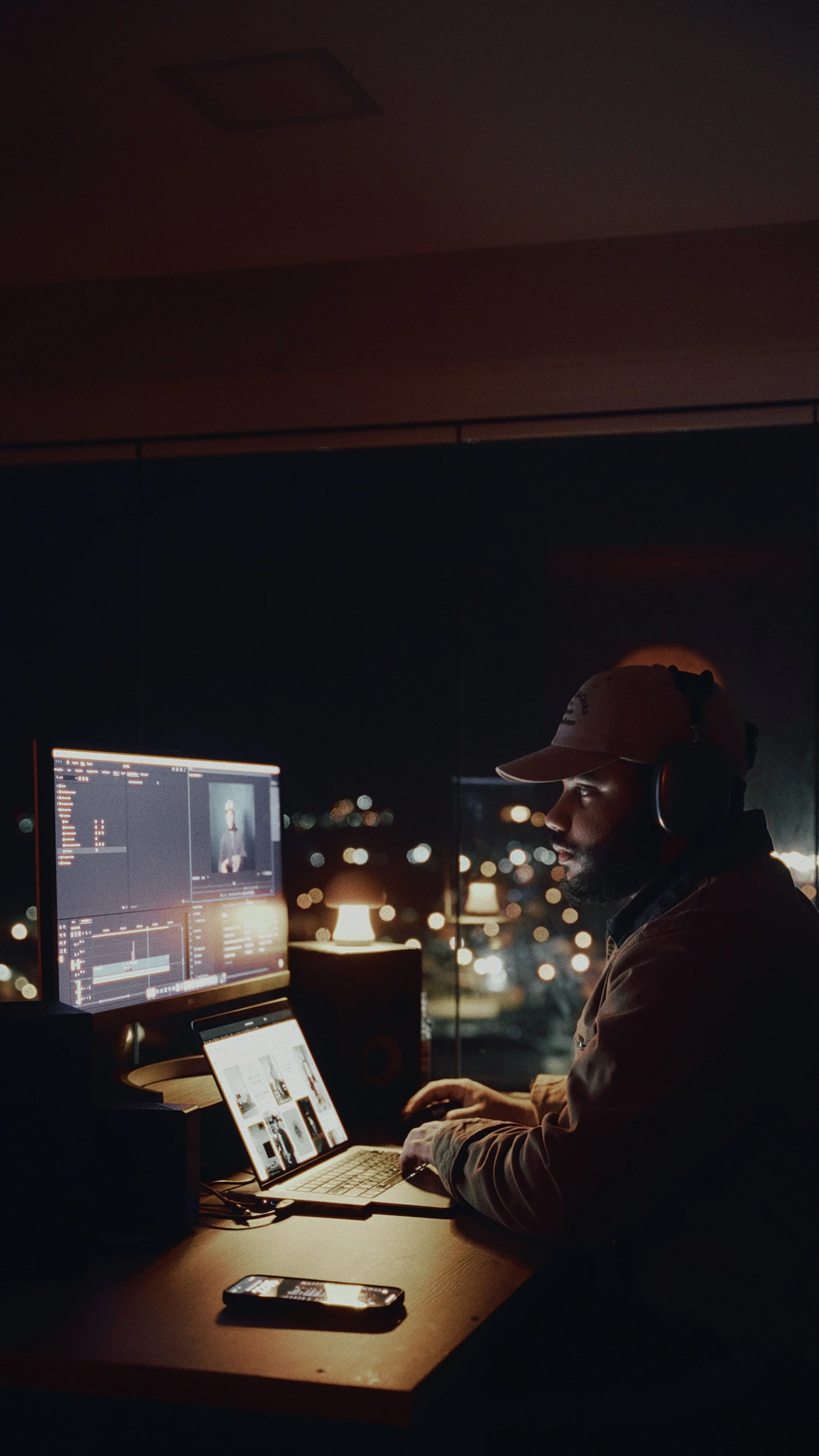 Homem trabalhando em edição de vídeo em um escritório com vista para a cidade à noite, usando fones de ouvido e duas telas de computador.