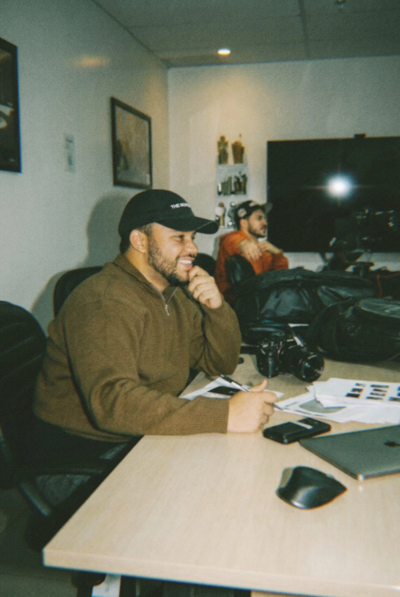 Homem sorrindo sentado em uma mesa de escritório com câmera, laptop, celular e papéis. Ao fundo, outra pessoa usando capacete e monitor de TV.