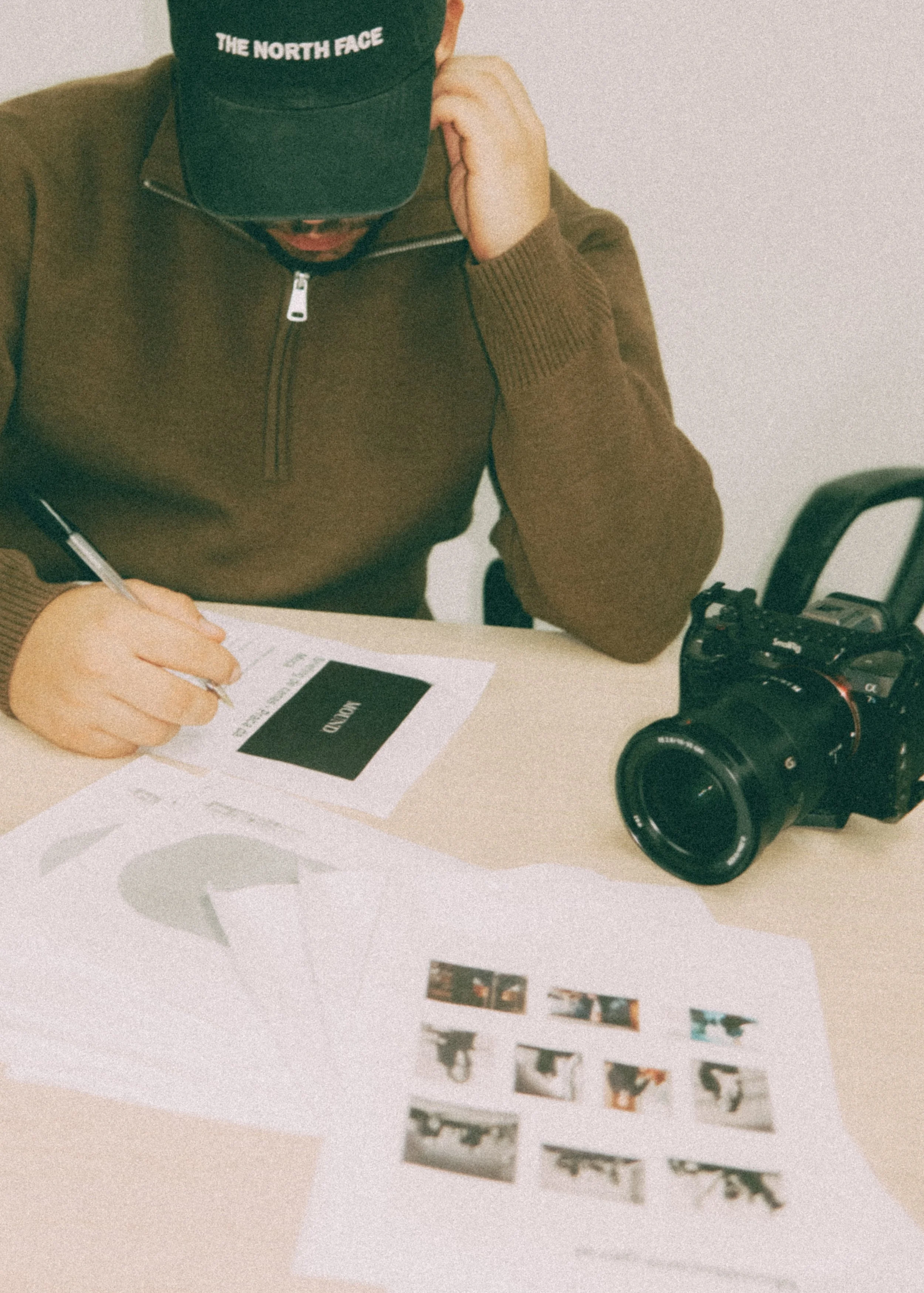 Pessoa de boné preto da The North Face, usando um suéter marrom, sentado à mesa, escrevendo em um caderno, com uma câmera profissional ao lado e fotos impressas na mesa.