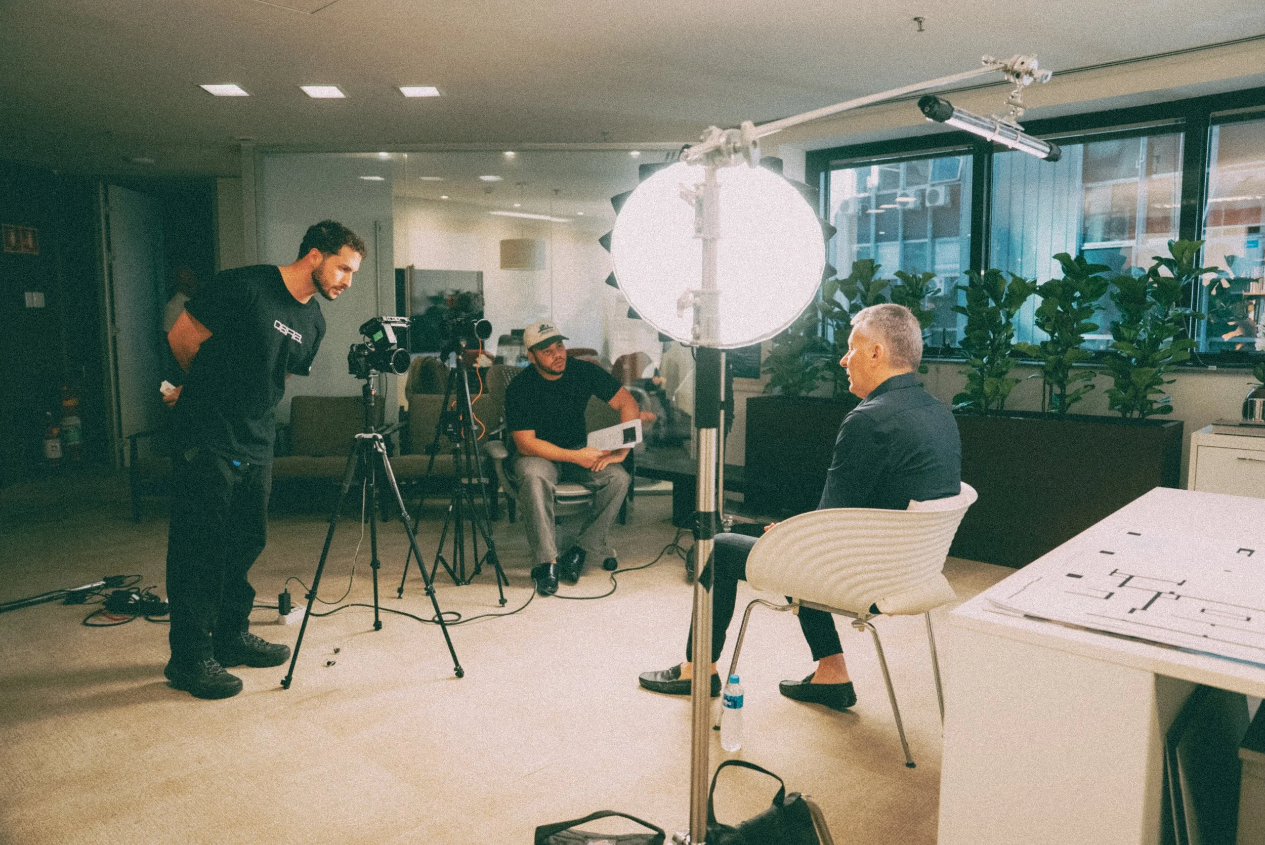 Cena de uma gravação de entrevista ou depoimento em um ambiente interno com três homens, duas câmeras, iluminação forte, uma pessoa segurando uma folha de papel e uma mesa com papelório ao lado.