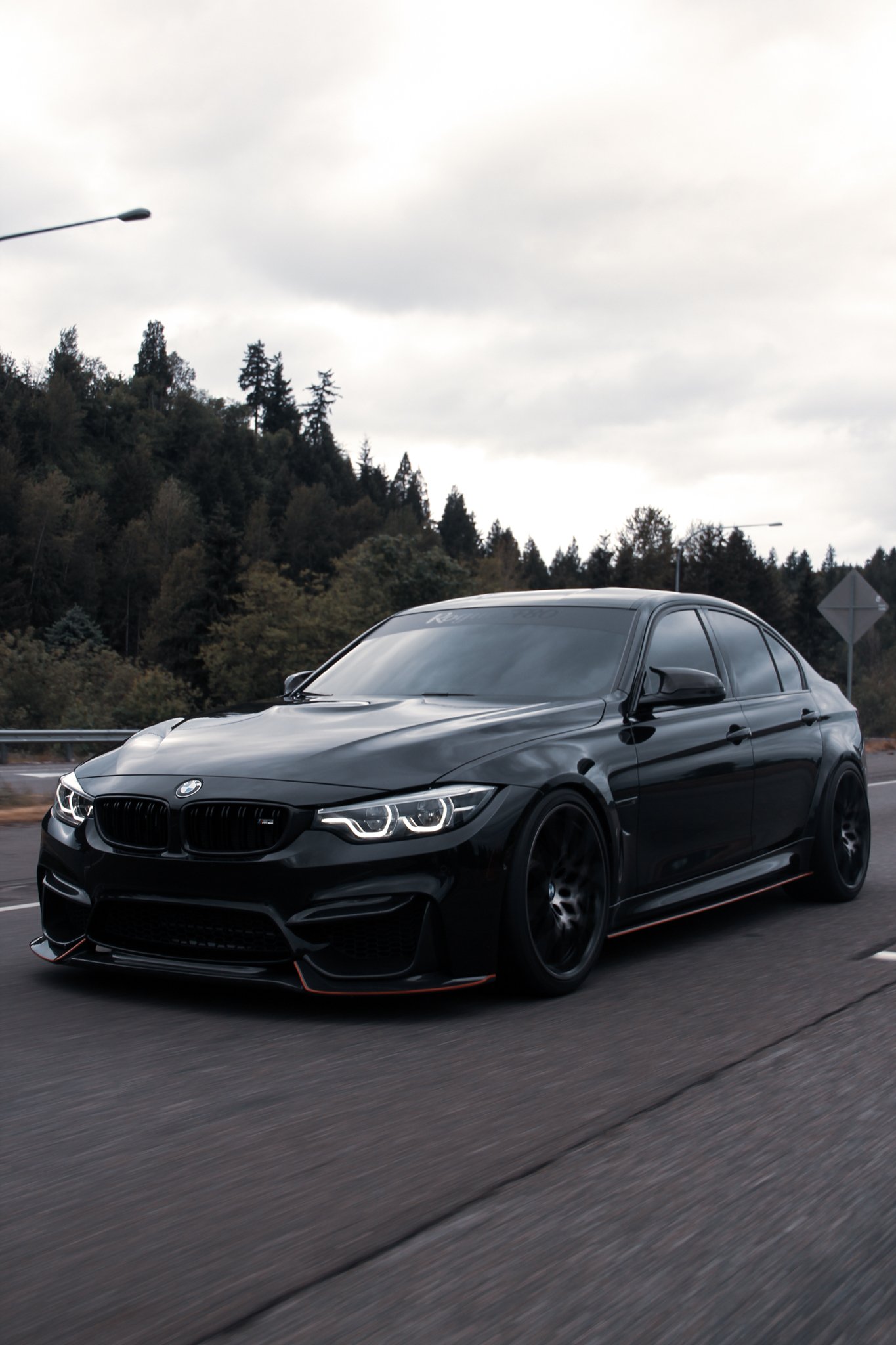 A black BMW sports car driving on a highway with trees and cloudy sky in the background.