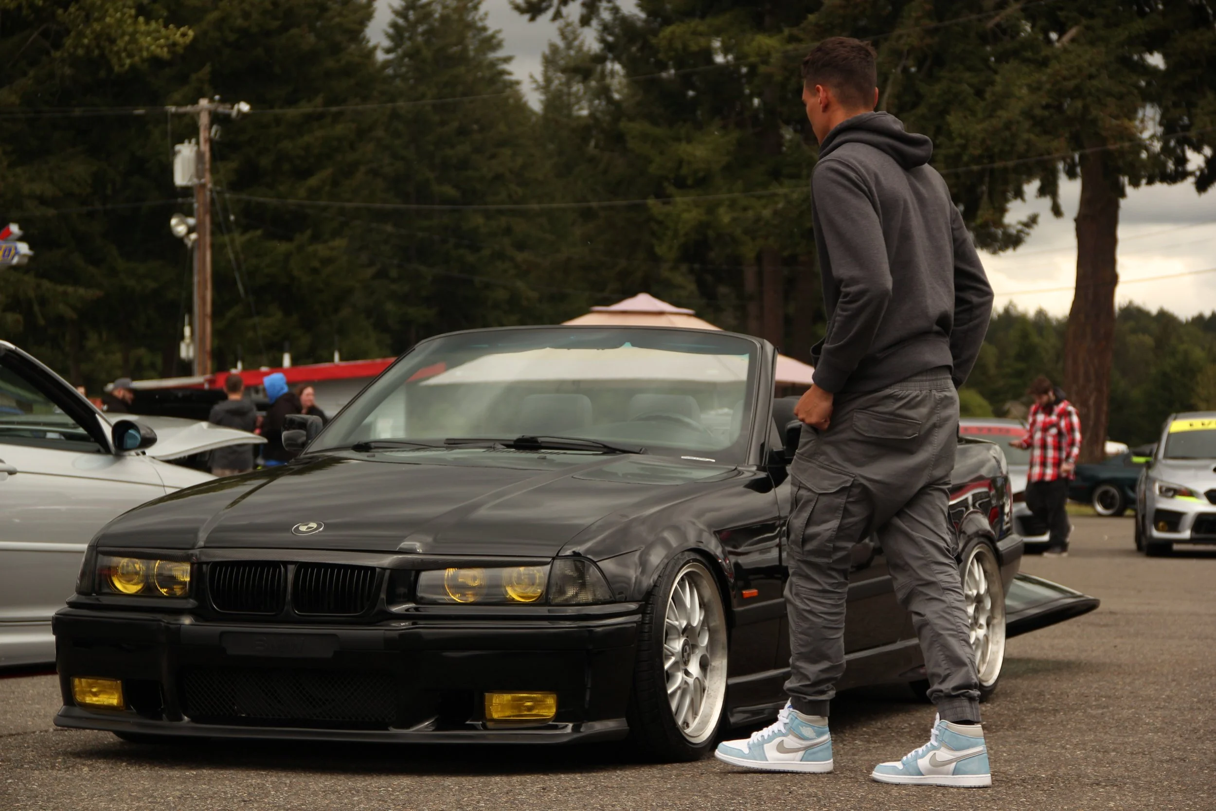 A man in gray hoodie and cargo pants looking at a black BMW convertible parked at a car meet or show, with other cars and people in the background.