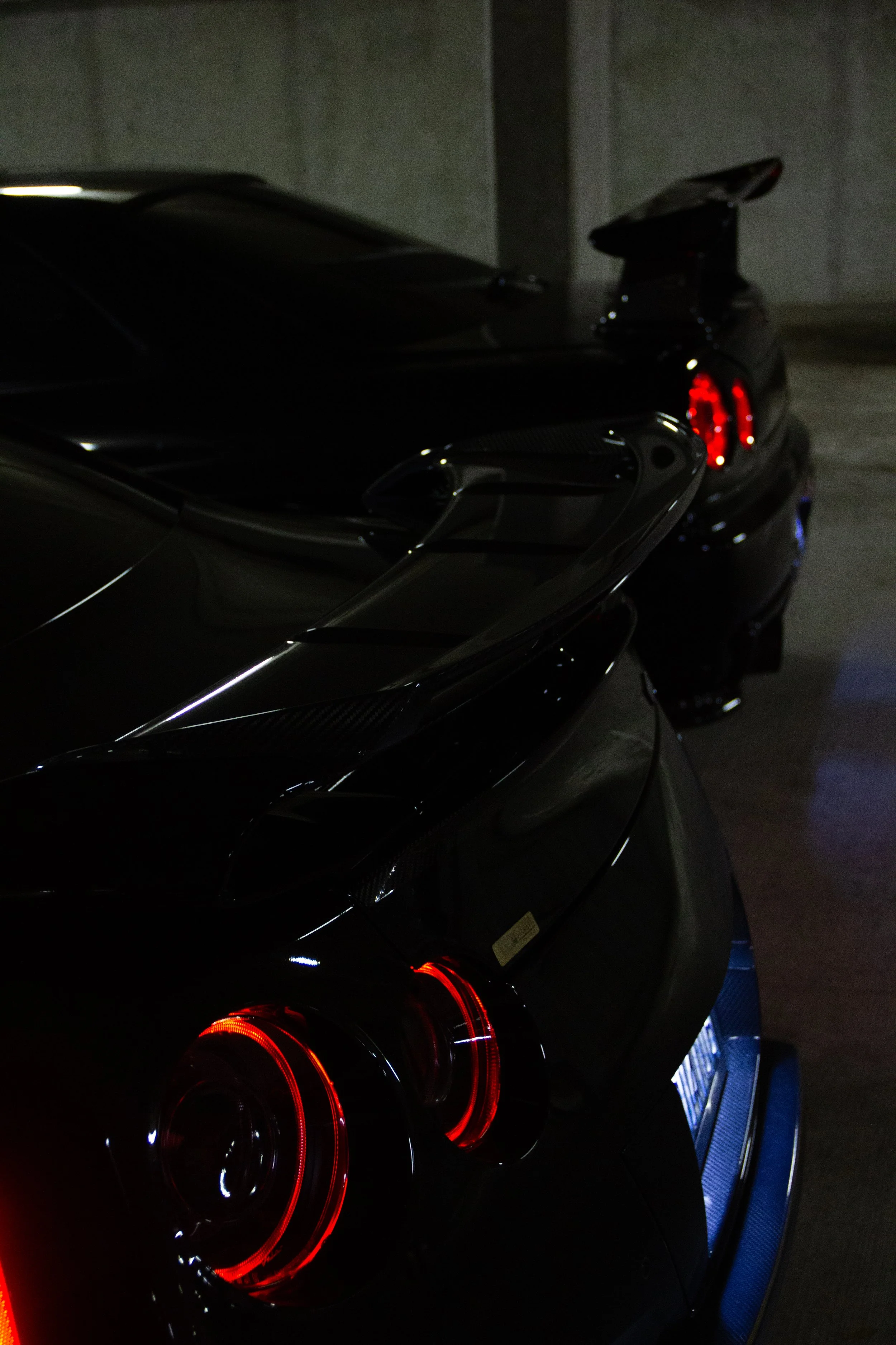 Close-up of the rear of a black sports car with illuminated circular red tail lights and a blue rear diffuser, parked in a dimly lit garage.