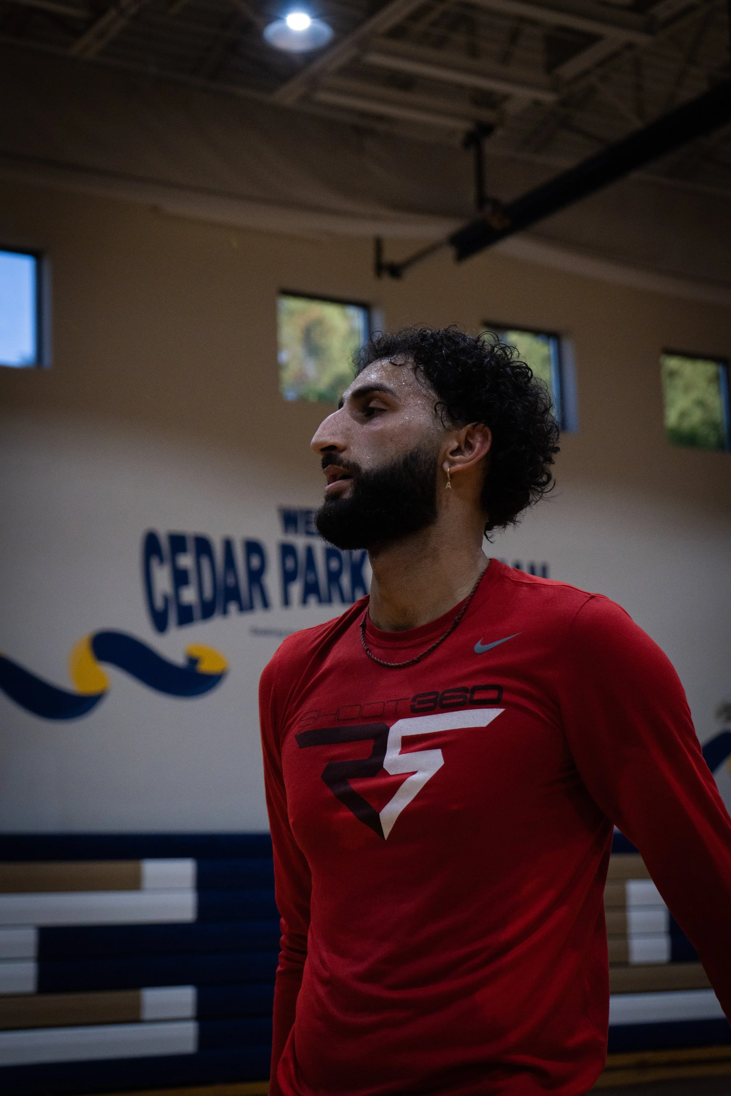 A man with curly dark hair and a beard, wearing a red athletic shirt and earrings, standing inside a gym with a sign that reads 'Cedar Park' behind him.