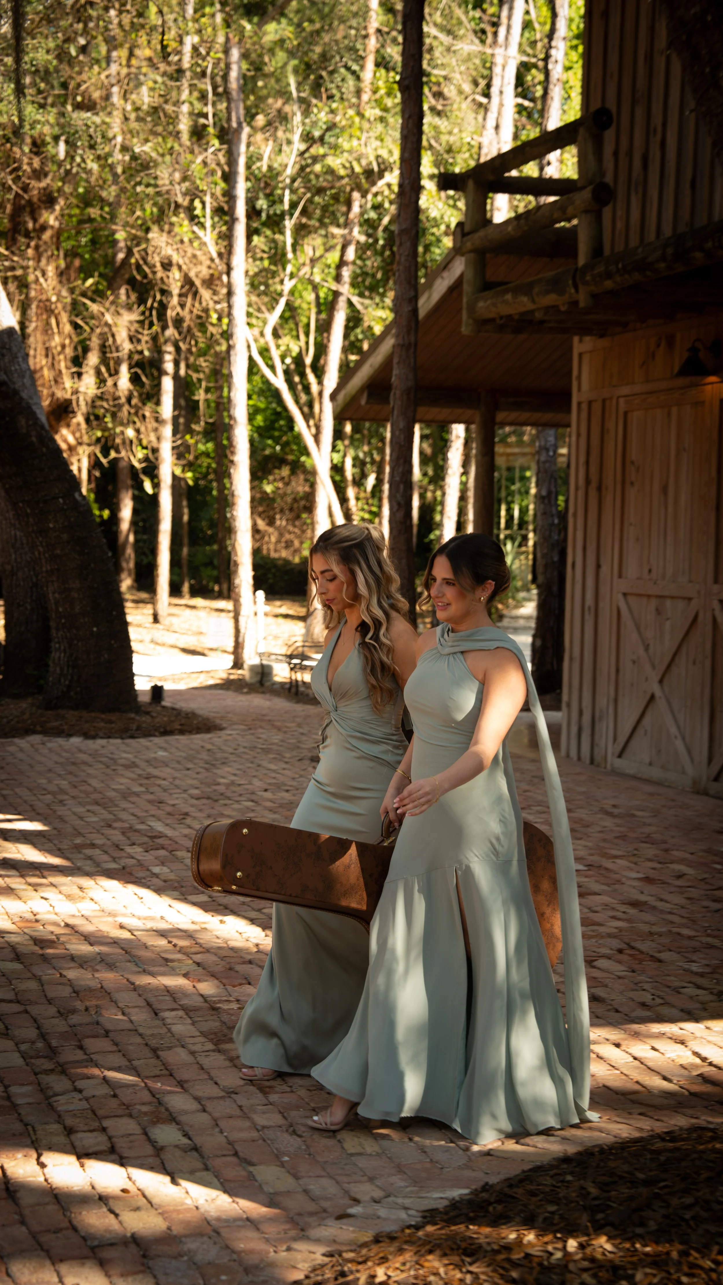 Two women in elegant dresses walking outdoors, one carrying a guitar case, with trees and a wooden building in the background.