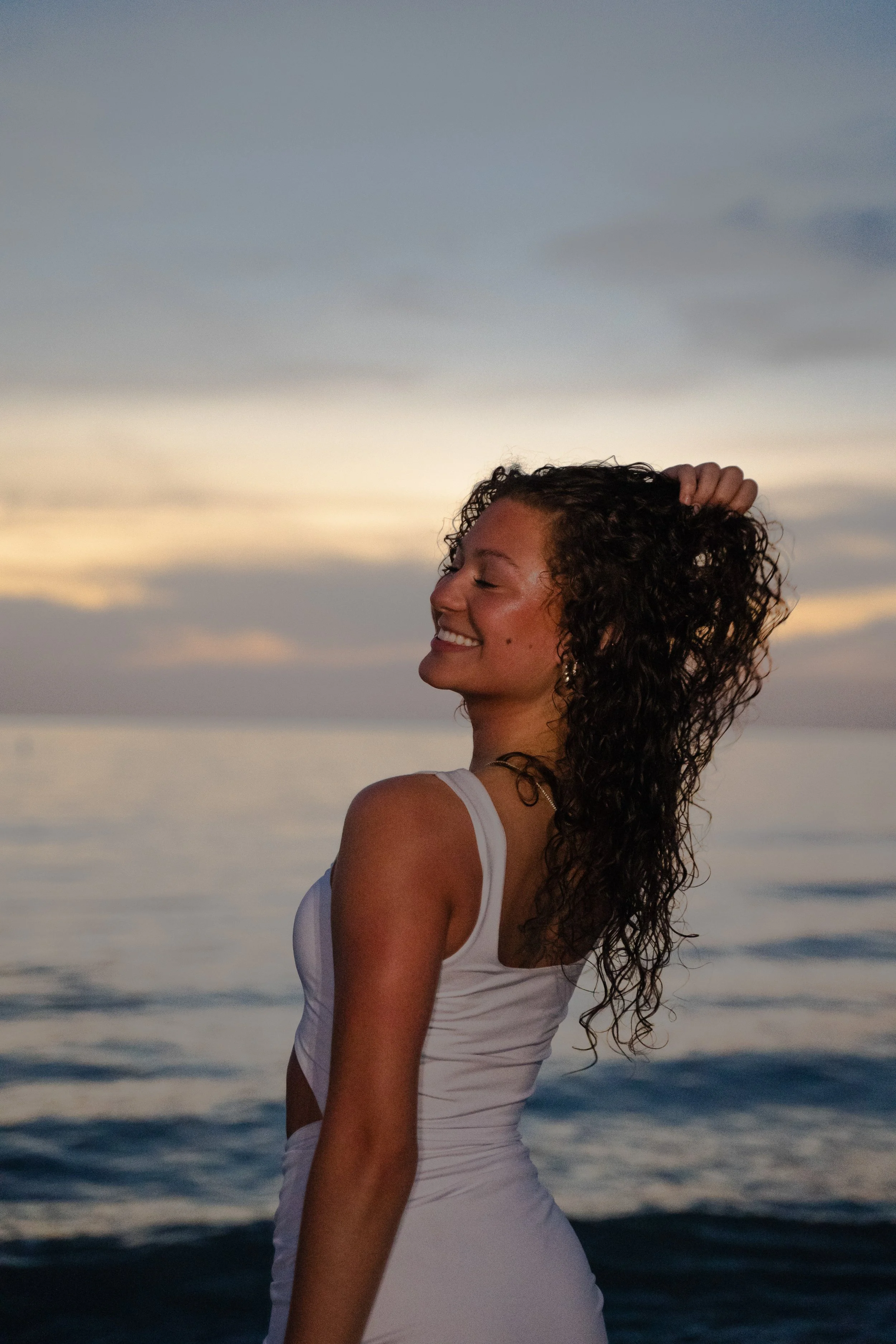 Young woman with curly hair standing near water at sunset, smiling with eyes closed, wearing a white sleeveless dress.