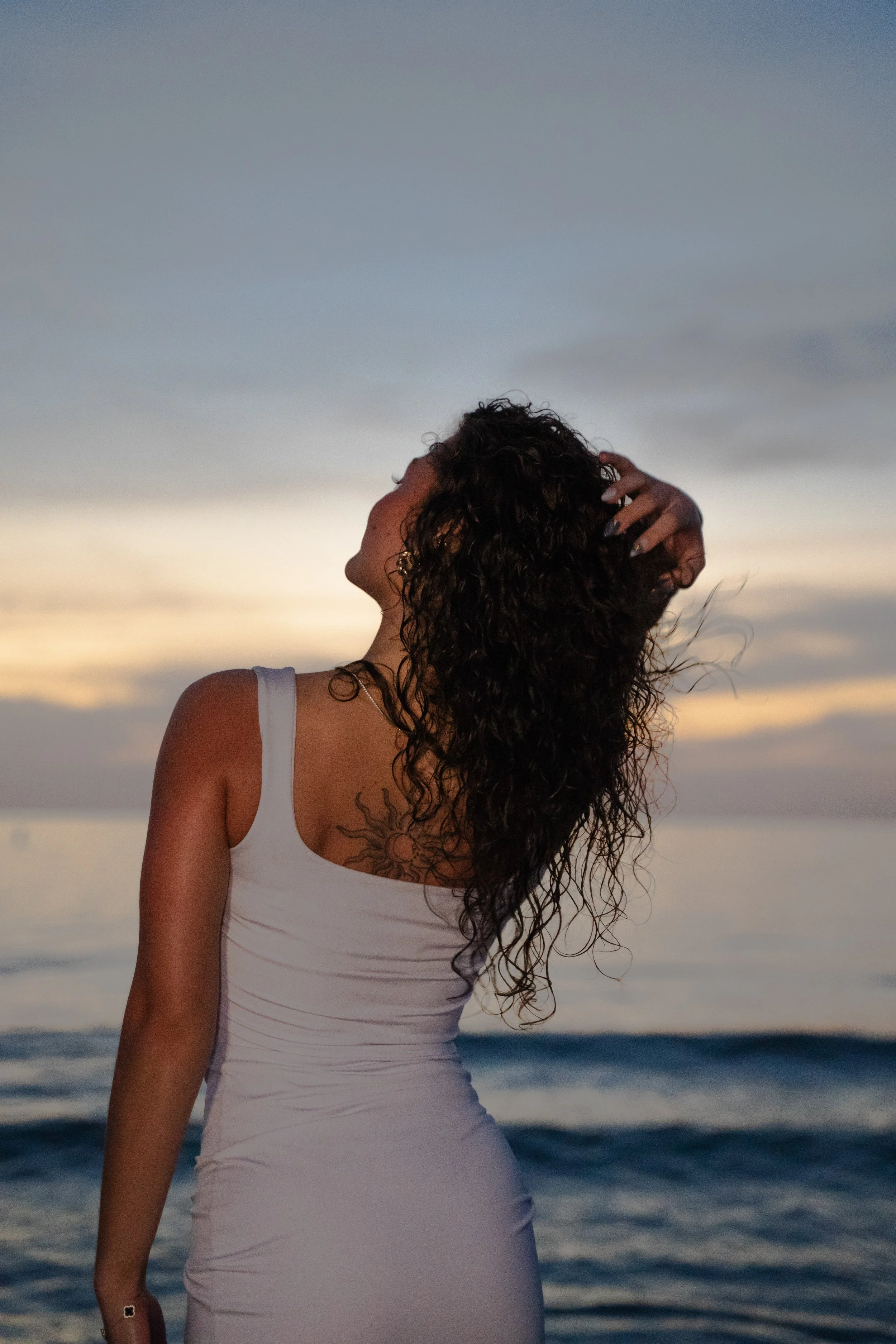A woman with curly hair wearing a white dress stands on a beach during sunset, with her head turned to the side and one hand in her hair.