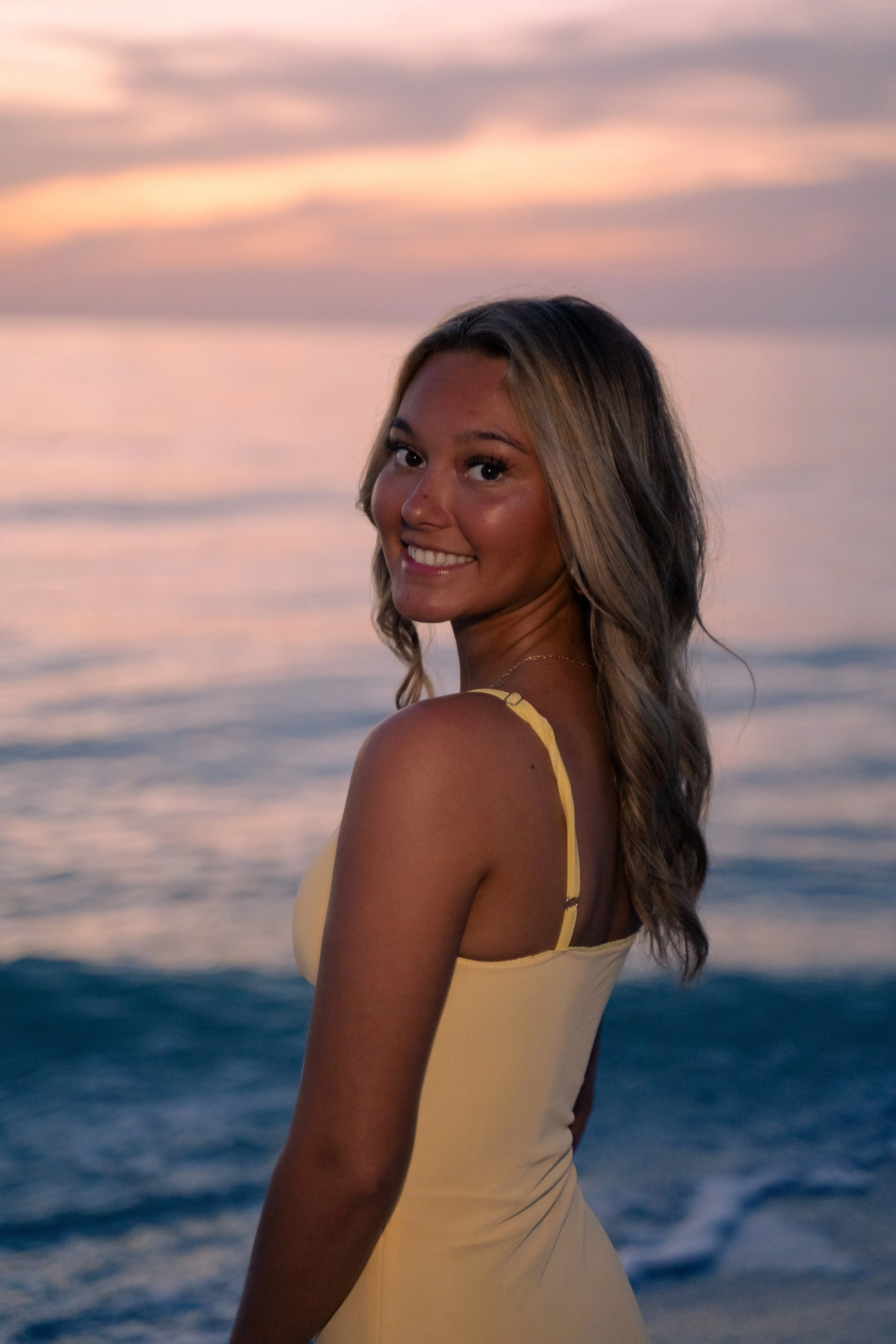 A young woman with shoulder-length wavy hair smiling at the camera at sunset near the ocean, wearing a yellow sleeveless dress.