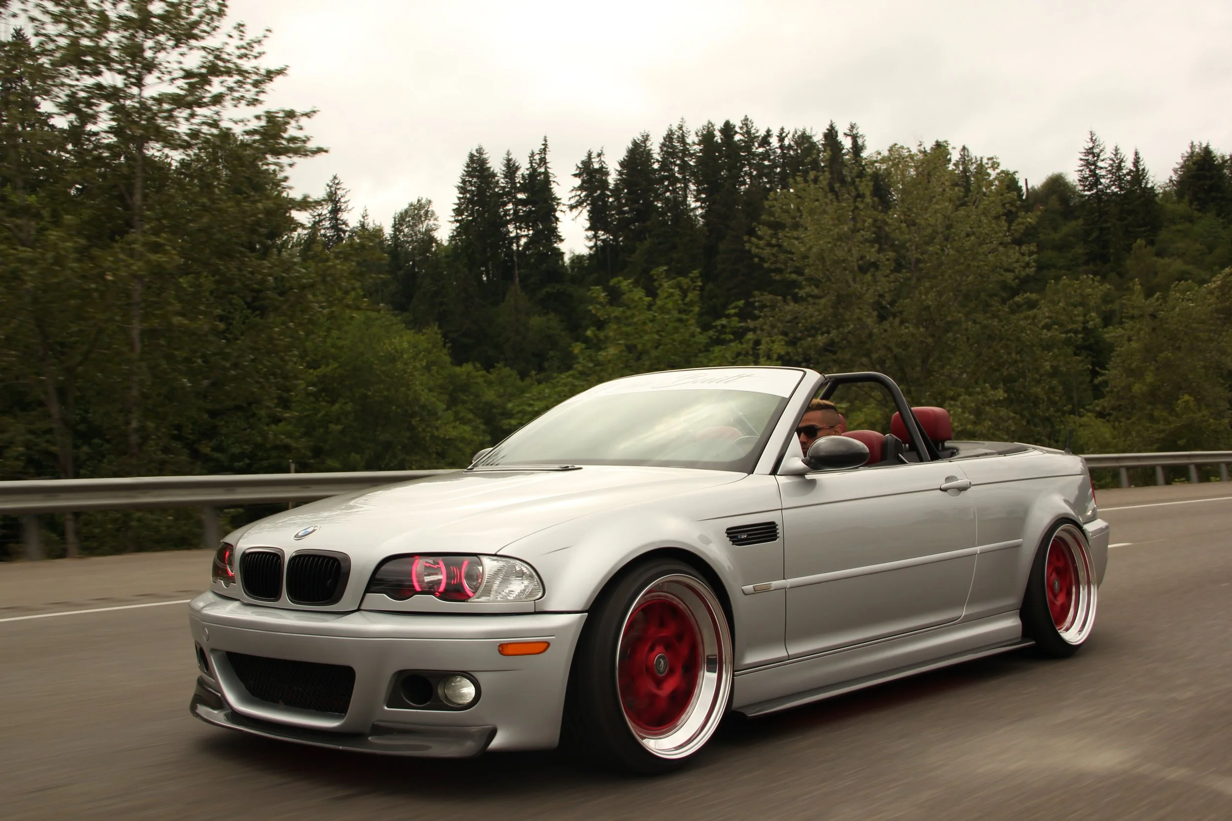 A silver BMW convertible with red accents on the wheels and seats, driving on a highway surrounded by trees.