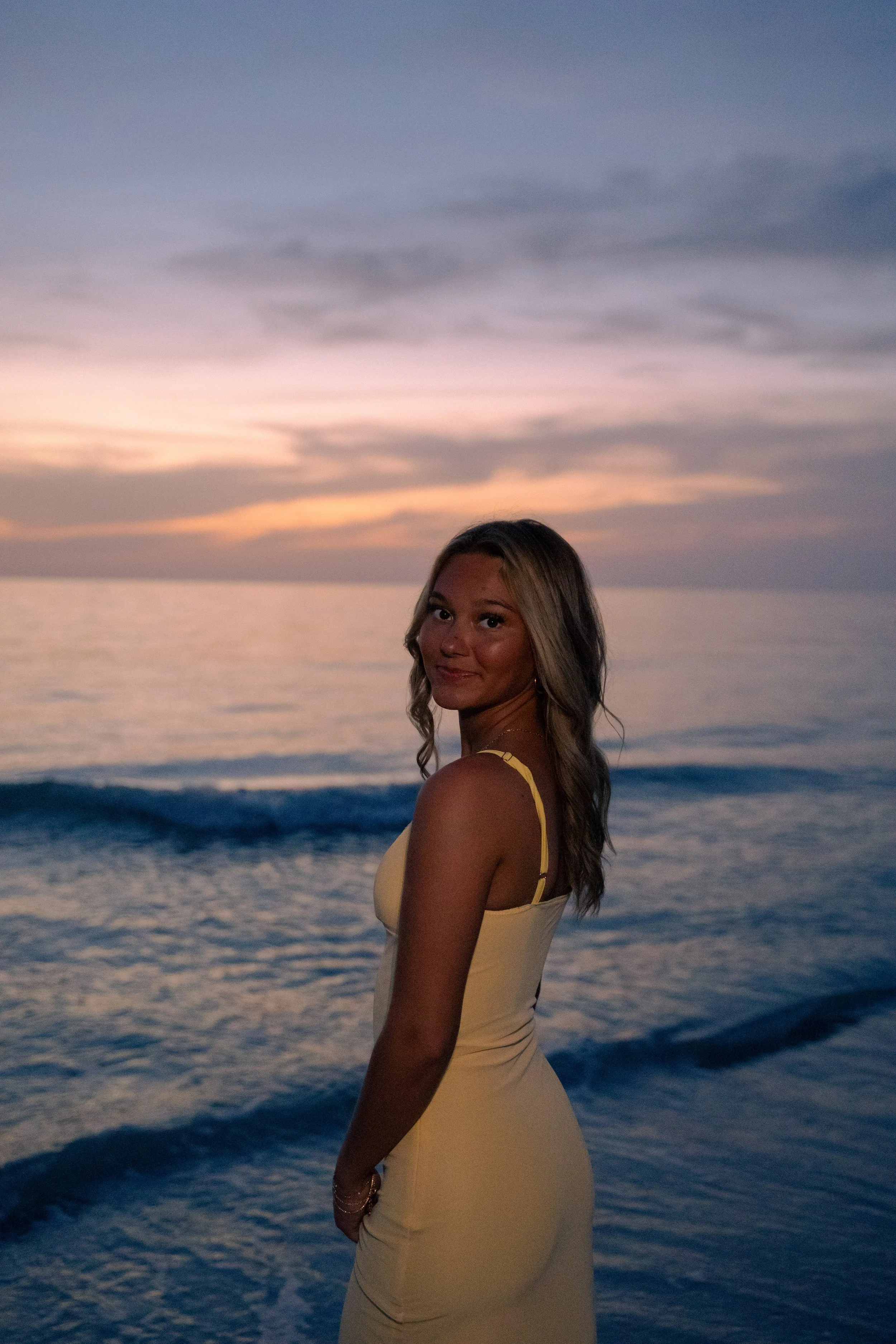 A woman in a yellow dress standing on the beach at sunset, facing sideways and smiling.