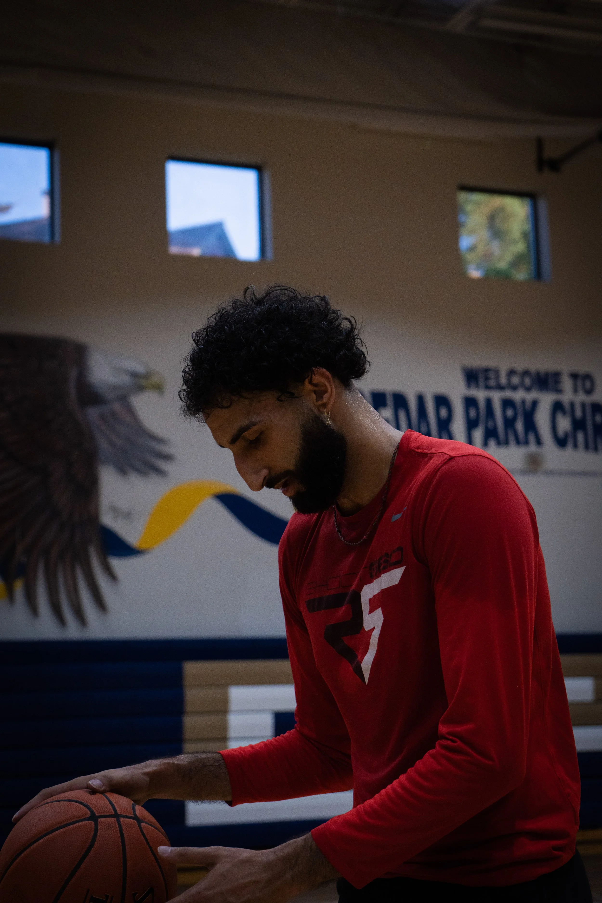 A young man with curly hair and a beard, wearing a red long-sleeve shirt, holding a basketball in a gym with a sign that says "Welcome to Cedar Park" and an eagle mural on the wall.