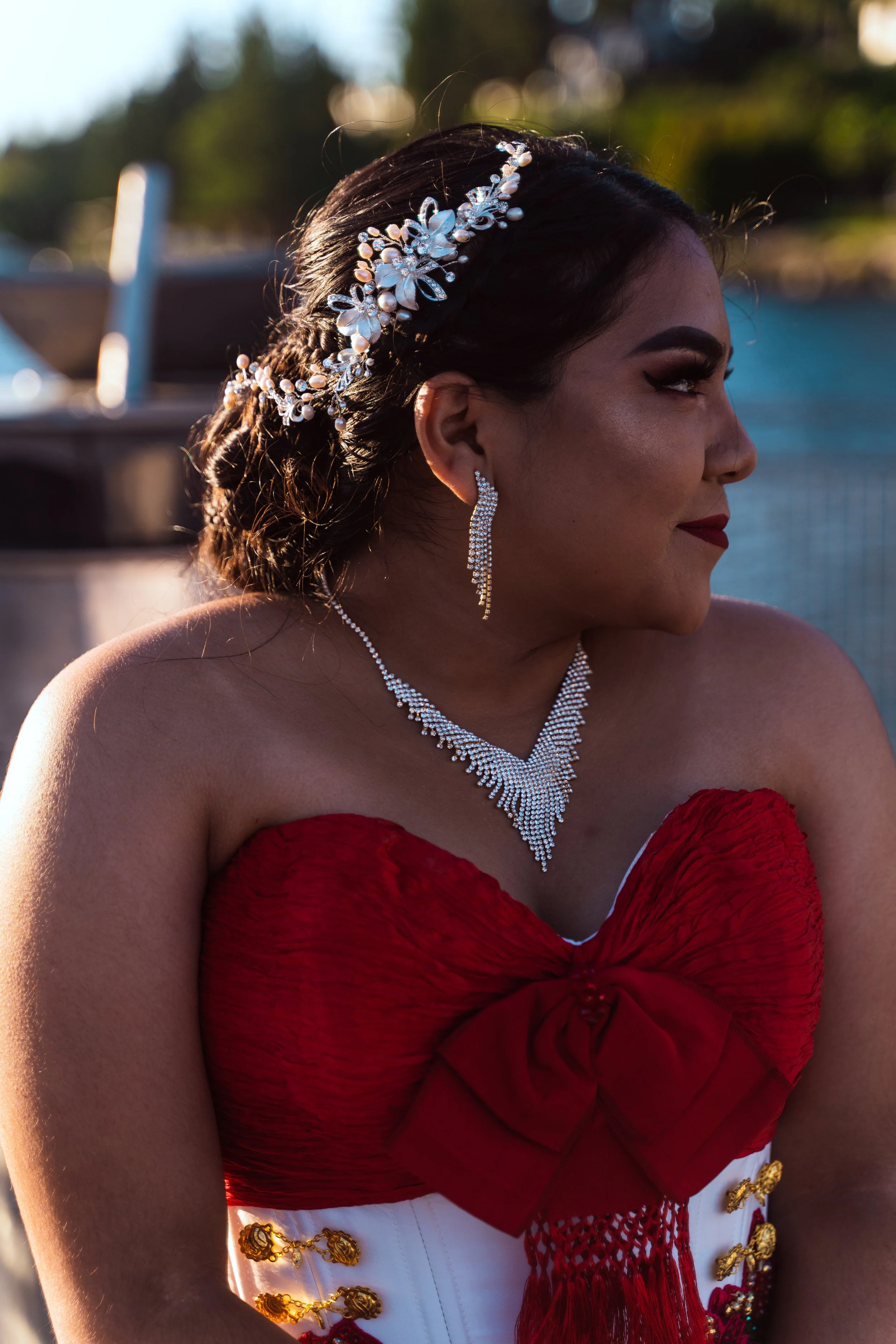 A woman dressed in red and white with jewelry, outdoors near water, with trees and blurred background.