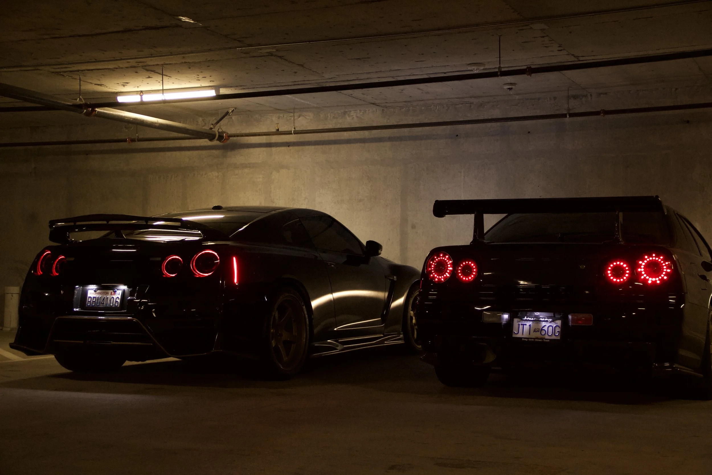 Two black sports cars with illuminated red taillights parked side by side in a dimly lit underground parking garage.