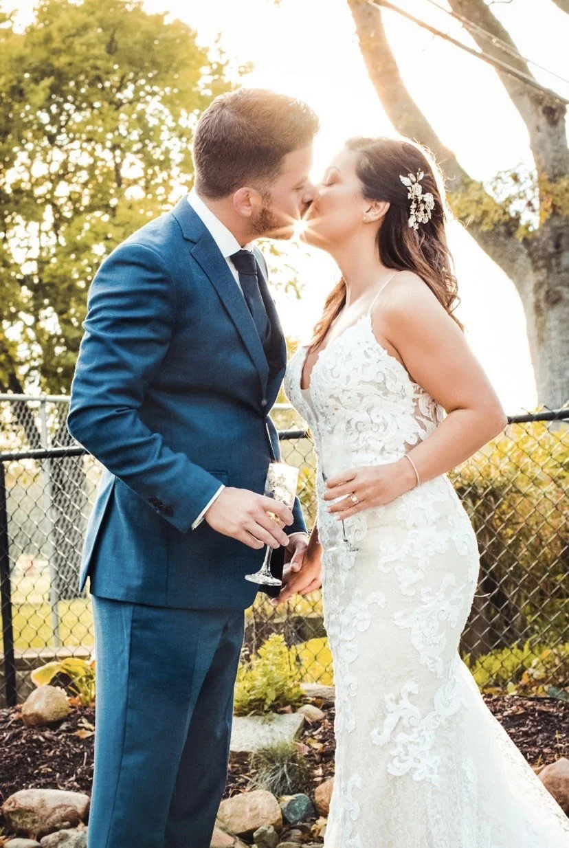 A newlywed couple sharing a kiss outdoors at sunset, with the groom in a blue suit holding a glass of wine, and the bride in a lace wedding dress with floral hair accessories, surrounded by trees and a chain-link fence.