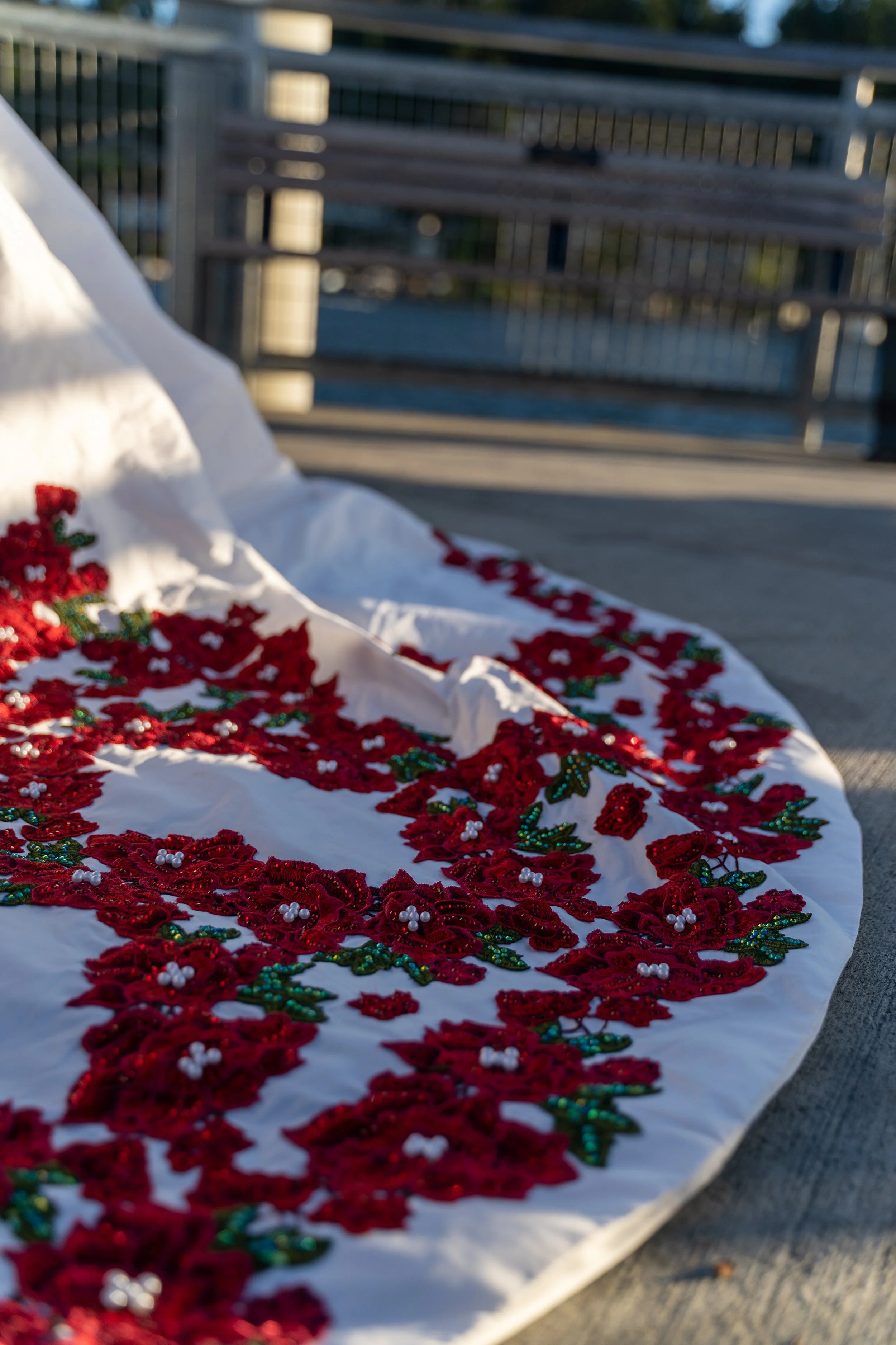 Close-up of a white embroidered fabric with red floral pattern and green leaves, lying on a flat surface outdoors with a blurred background.