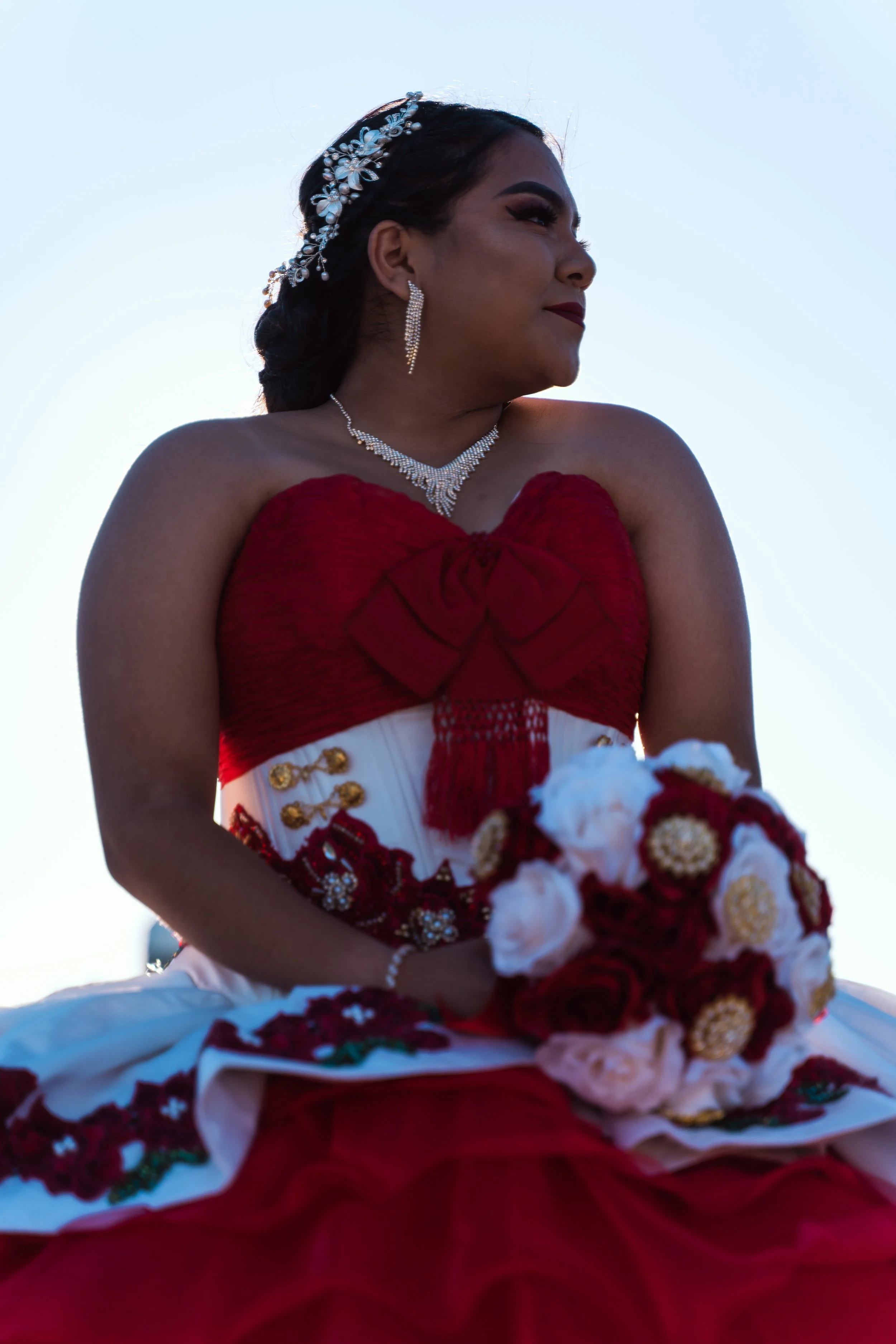 Woman in a red and white dress holding a bouquet of red and white flowers, wearing jewelry, and posing against a clear blue sky.