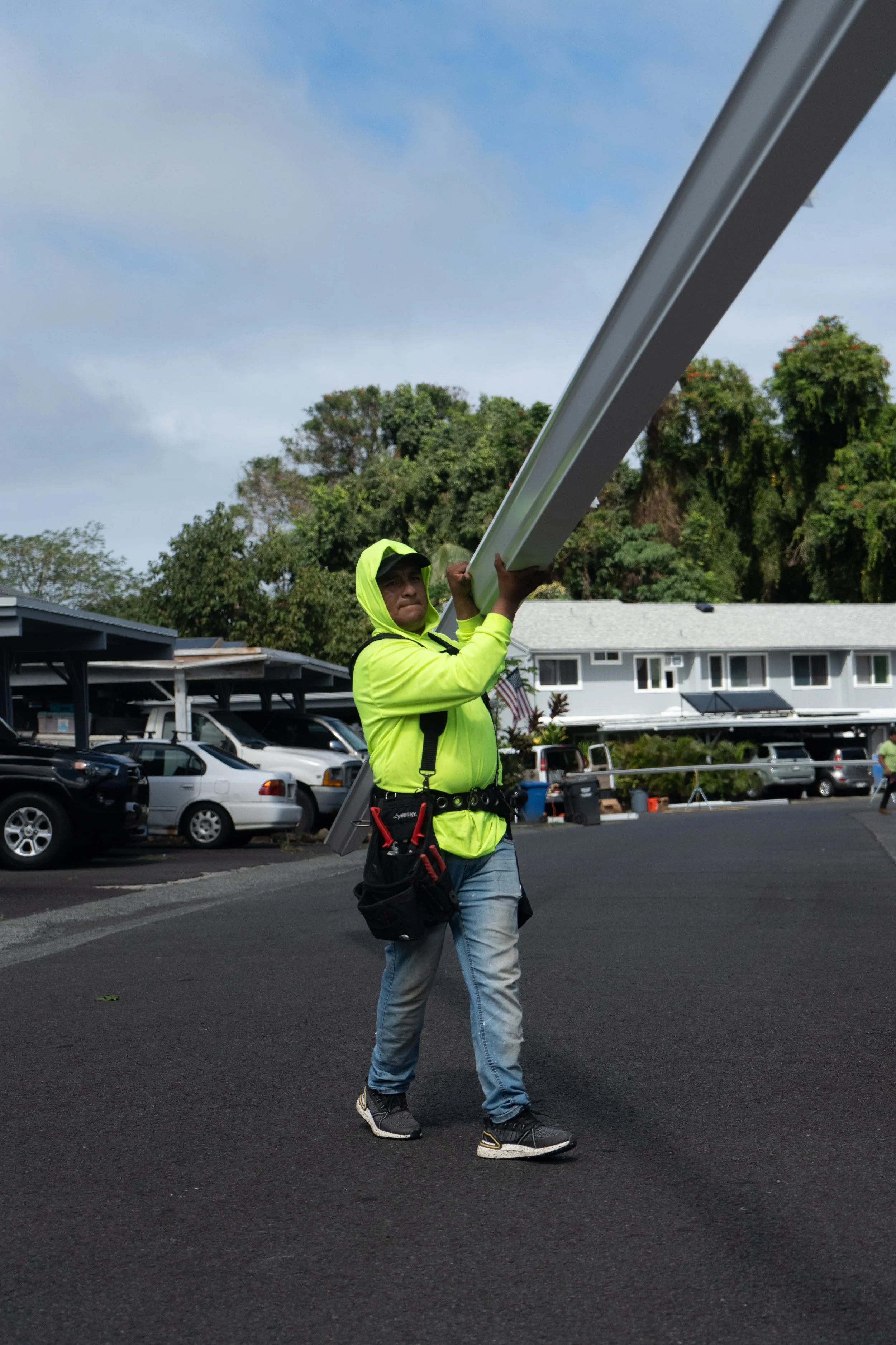 A man in a neon yellow hoodie and jeans carries a long white pole in a parking lot with parked cars and trees in the background.