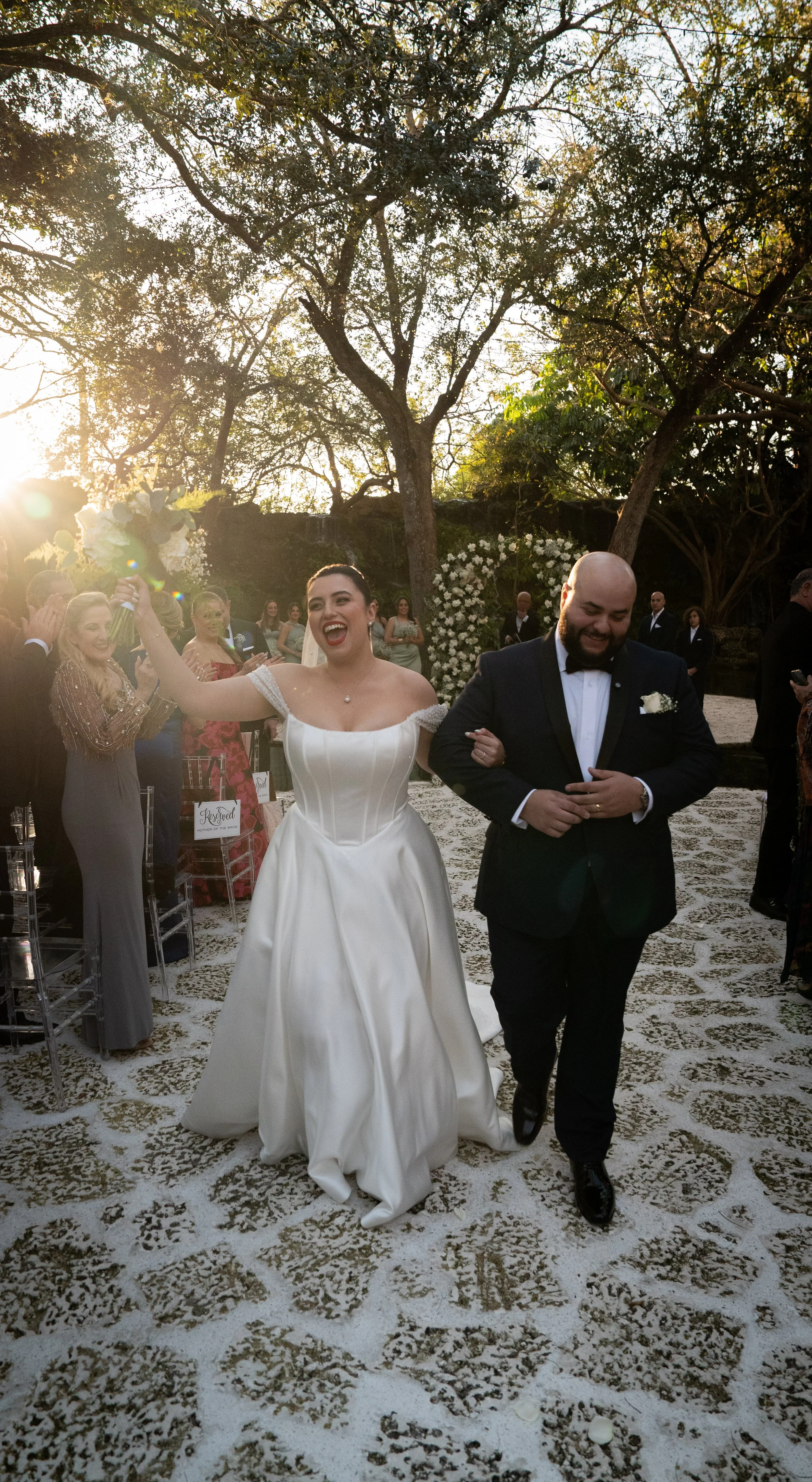 Bride in a white wedding gown holding a bouquet, walking down the aisle with a joyful expression, arm linked with a groom in a black tuxedo, at an outdoor wedding ceremony during golden hour.